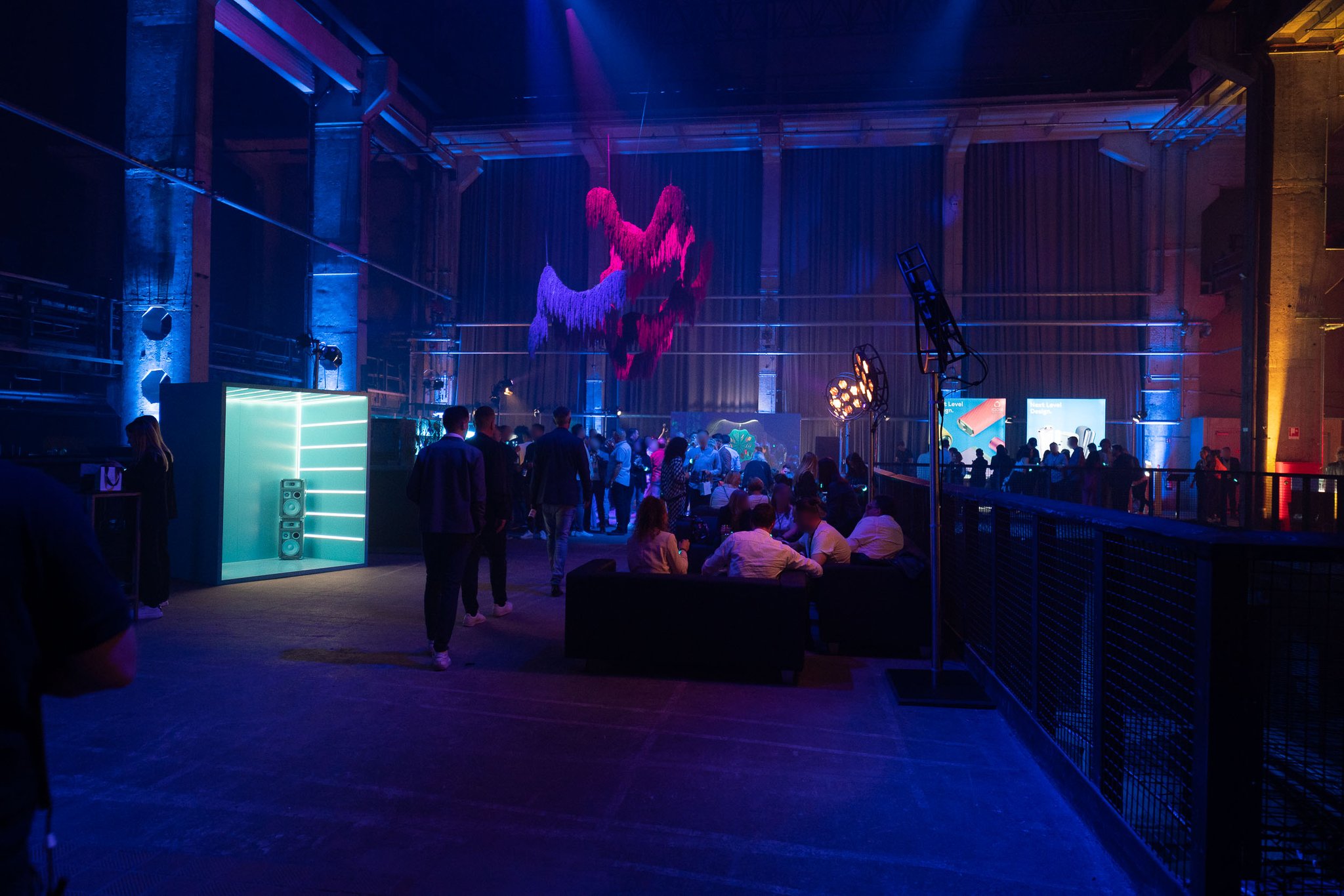 A lounge area with low seating and guests under blue light, with a red hanging sculpture and teal-lit booths in an industrial hall.