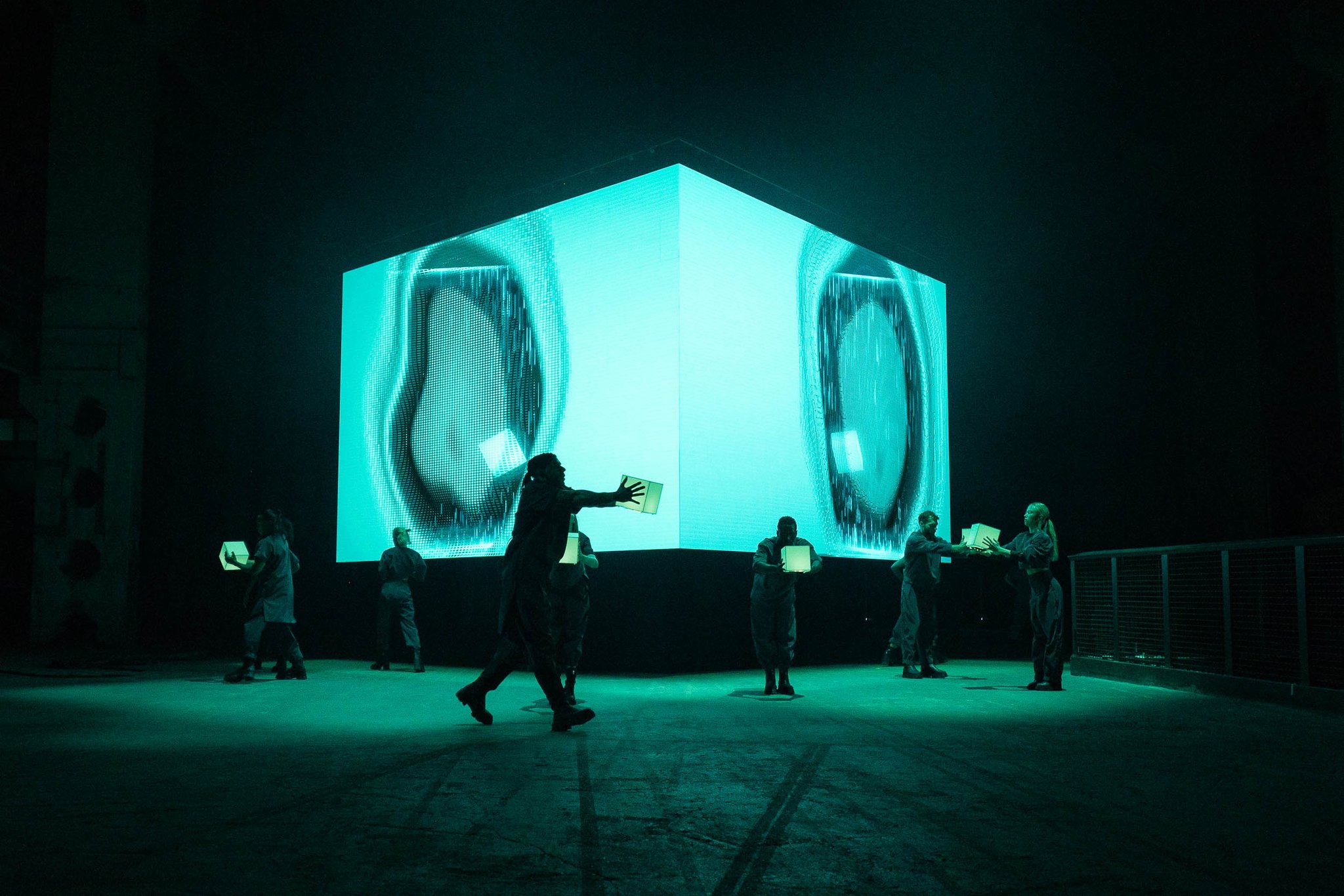 A large glowing cube with projected imagery stands in the middle of a dark hall, with performers moving around it.