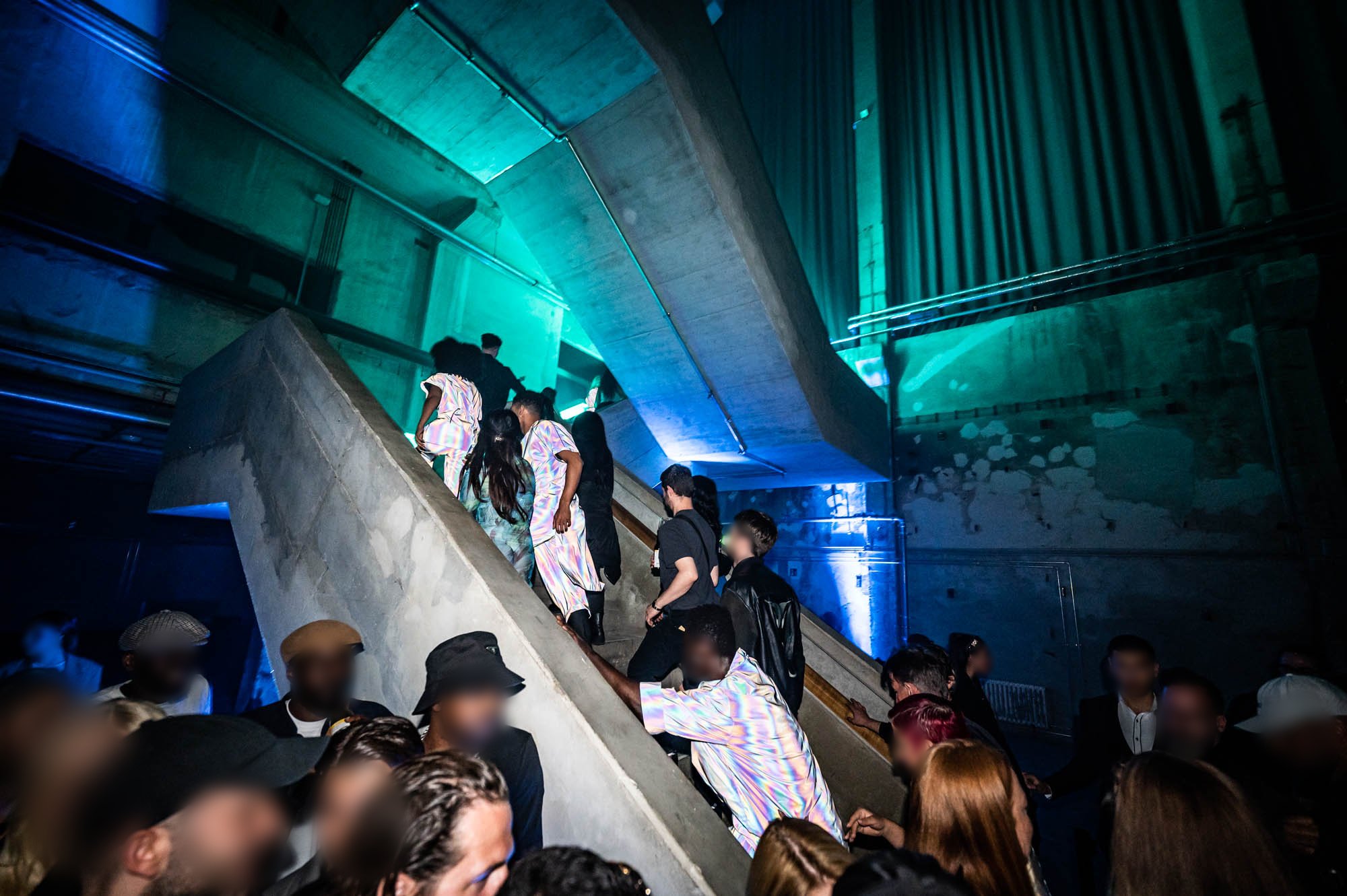 A crowd of guests climbs a wide concrete staircase under teal and blue light in an industrial hall.