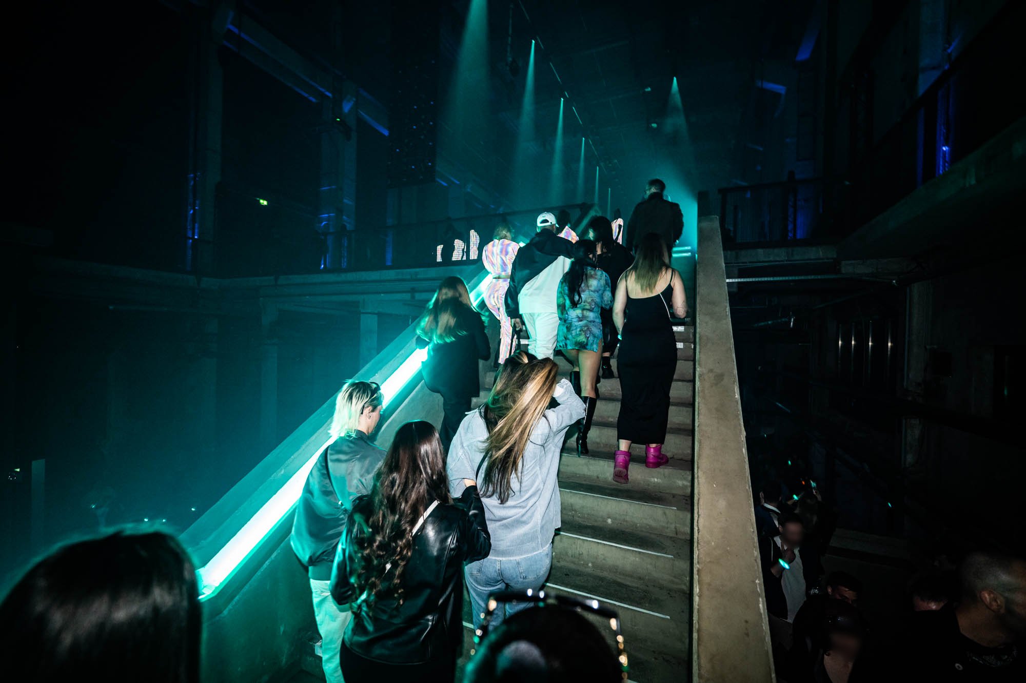 Guests walk down a long concrete staircase lit by teal light strips inside an industrial building.