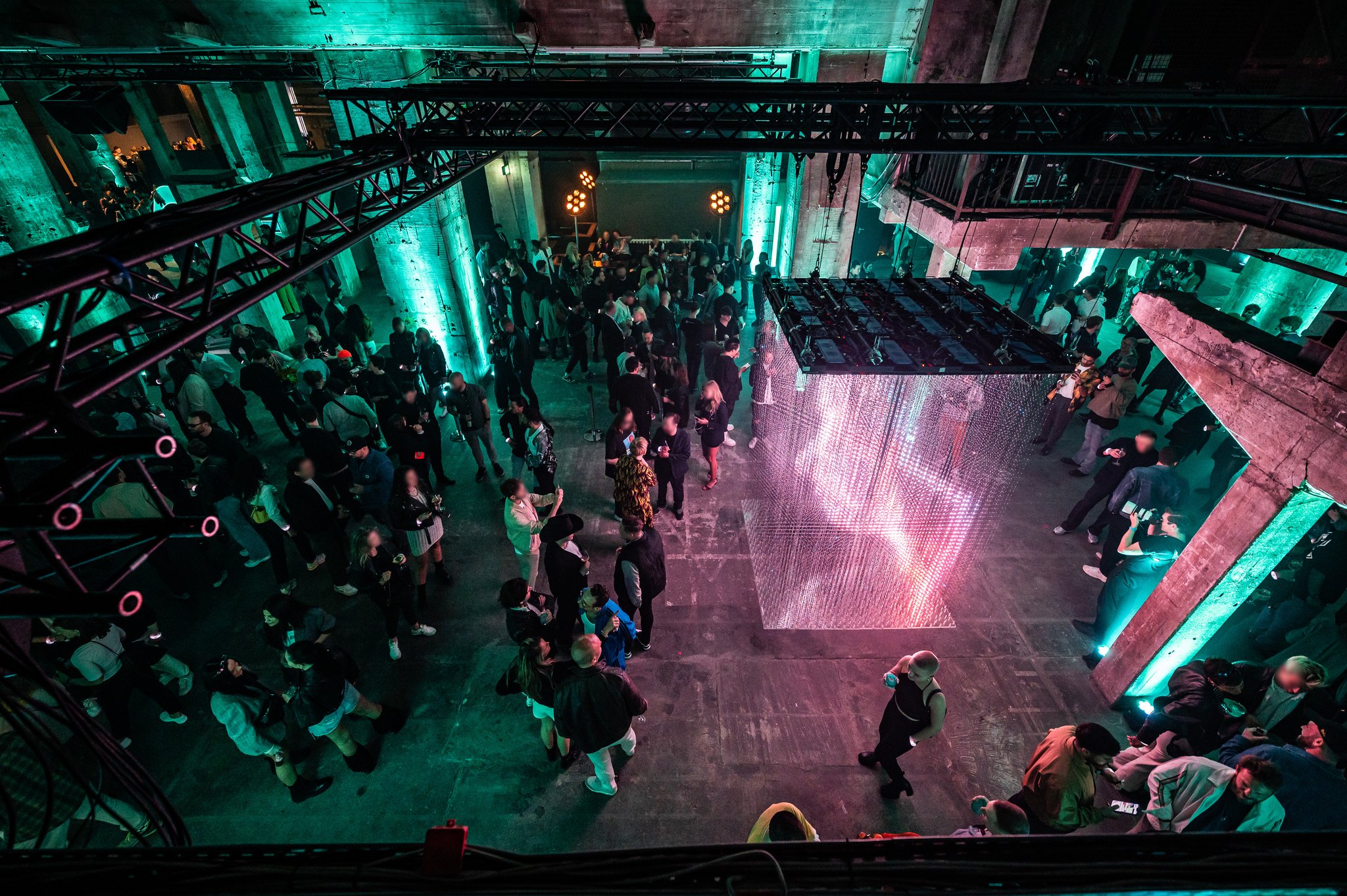 An overhead view of a crowd surrounding a large floor projection of pink light shapes, framed by teal-lit steel structures.