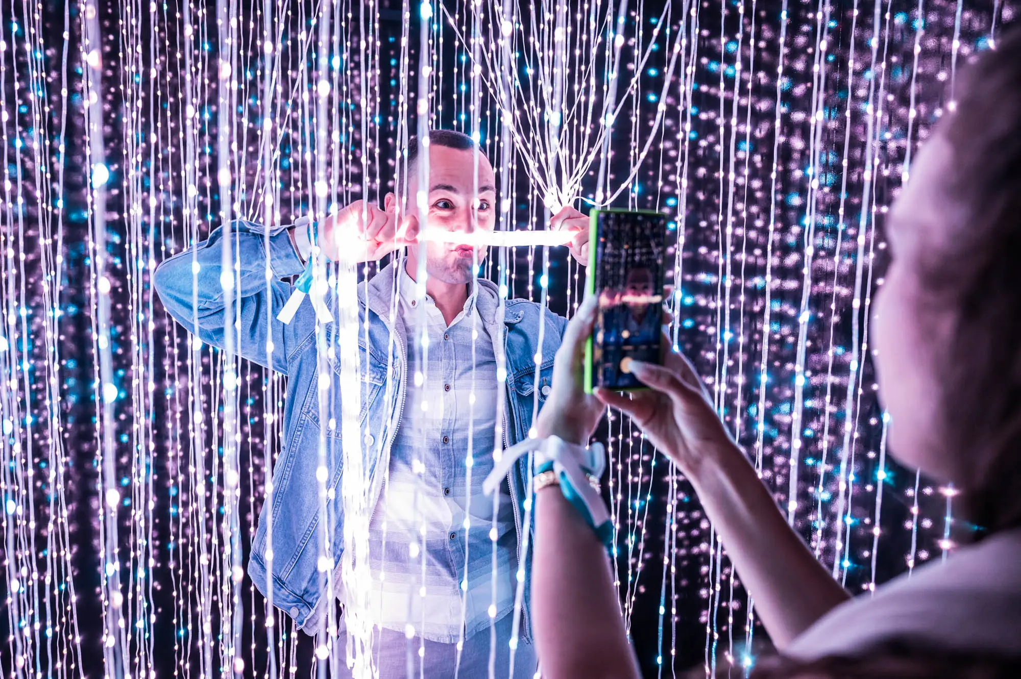 A man playfully poses with glowing lights while someone takes his photo in a room filled with hanging LED strands. BattleRoyalStudios