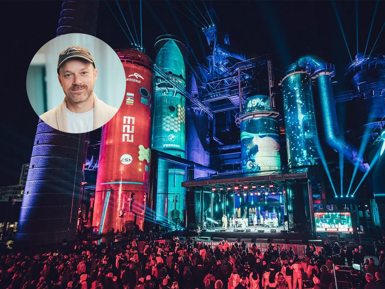 A futuristic event with large, illuminated industrial structures, featuring a crowd and a stage, with an inset portrait of Brendan Shelper in the top left corner. Battle Royal Studios