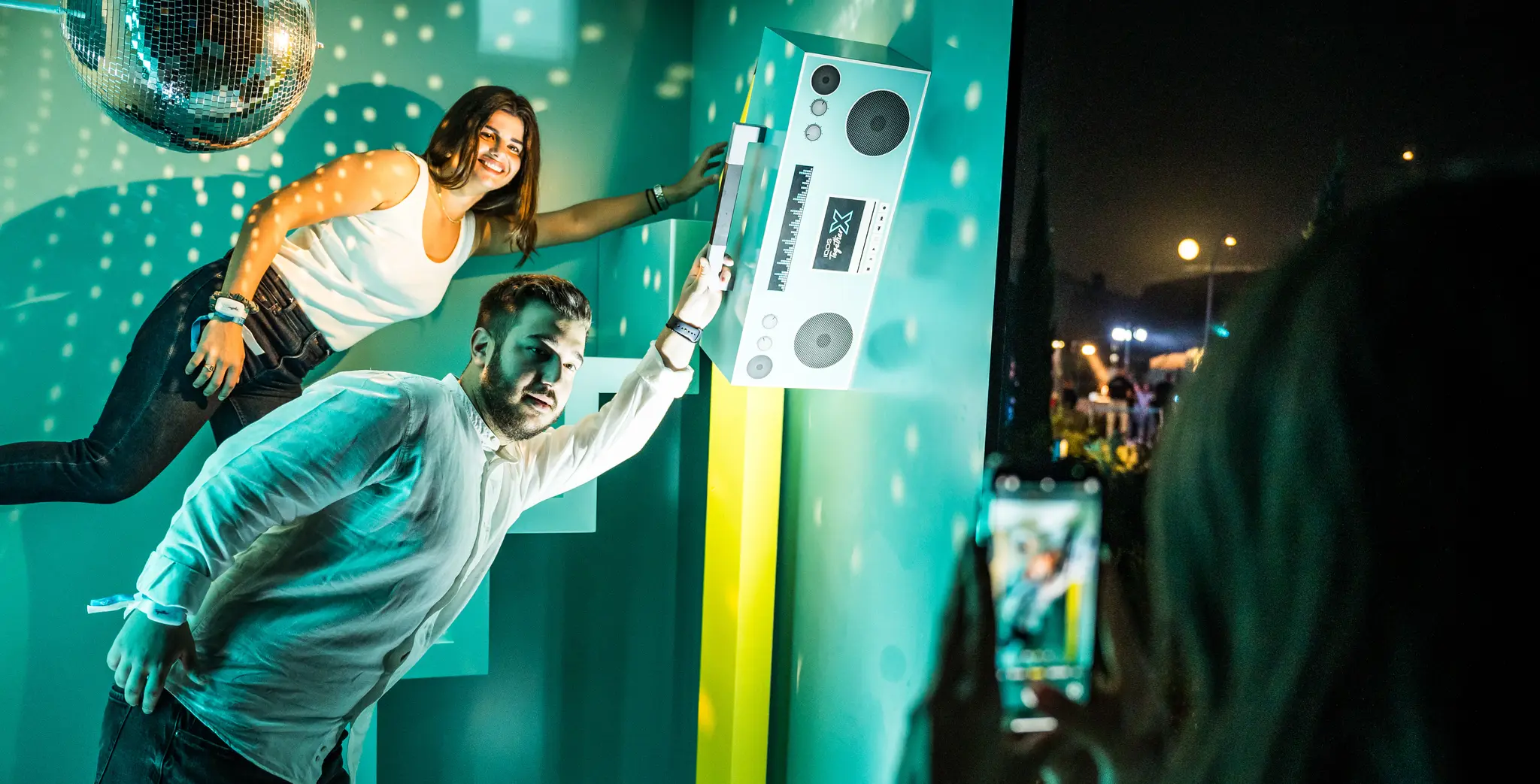 A man and woman pose playfully next to a giant boombox under a disco ball, while someone takes a photo with their phone. Battle Royal Studios