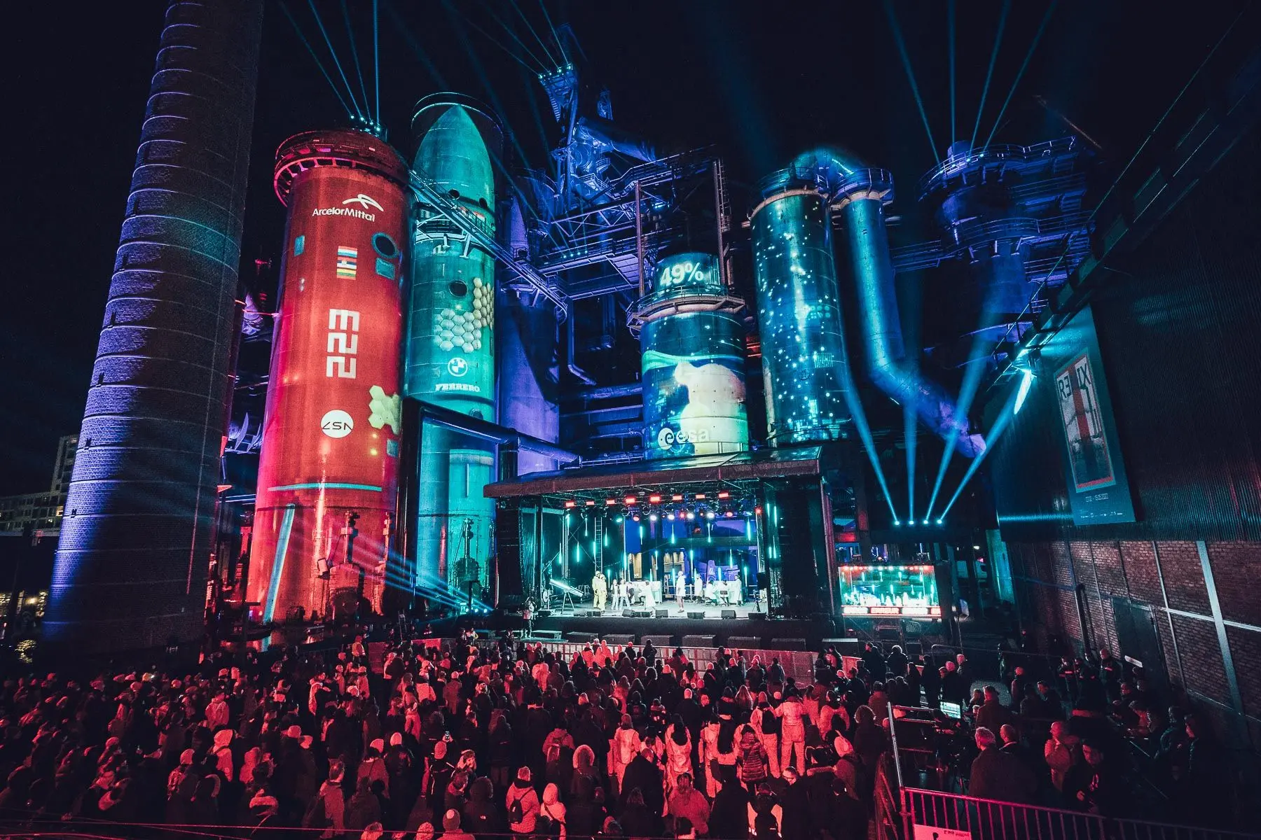 A crowd gathers in front of a lit industrial stage with colorful projections. Battle Royal Studios