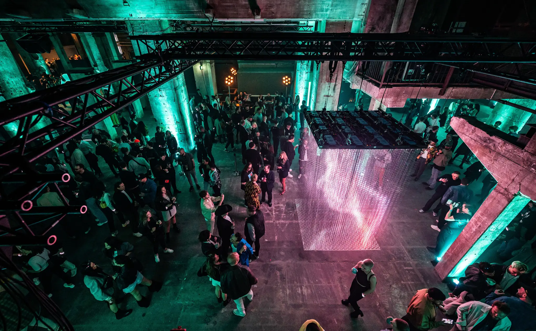 An overhead view of a crowd mingling in an industrial-style venue with colorful lighting and a digital art installation. Battle Royal Studios