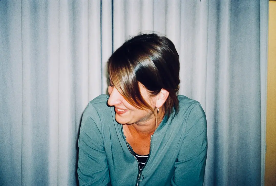 Judith Hoch in a teal top smiles while looking away, standing in front of gray curtains. Battle Royal Studios