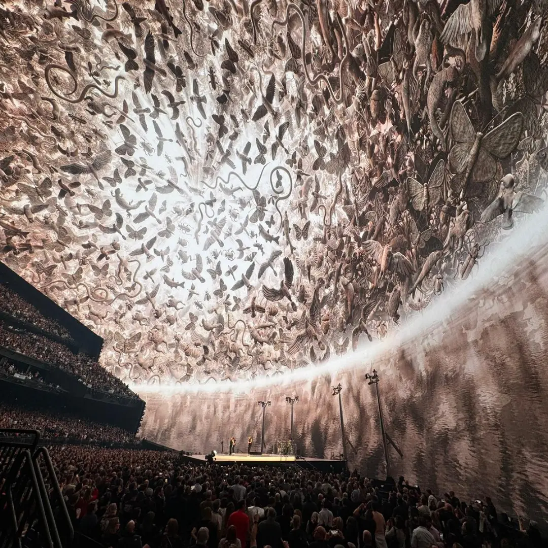 A vast audience watches a performance under a large dome covered with intricate artwork featuring birds, butterflies, and other creatures swirling toward a bright center. Battle Royal Studios