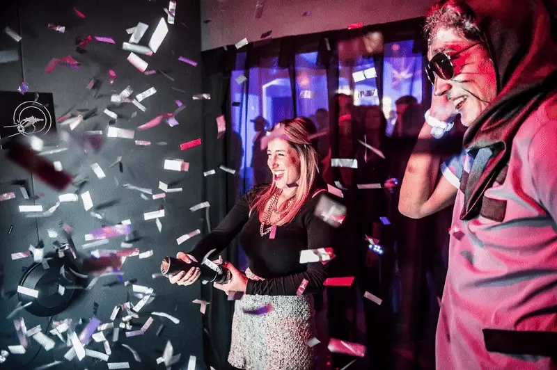 A woman joyfully sprays confetti from a bottle, surrounded by scattered pieces of paper as she celebrates in a lively atmosphere. Battle Royal Studios