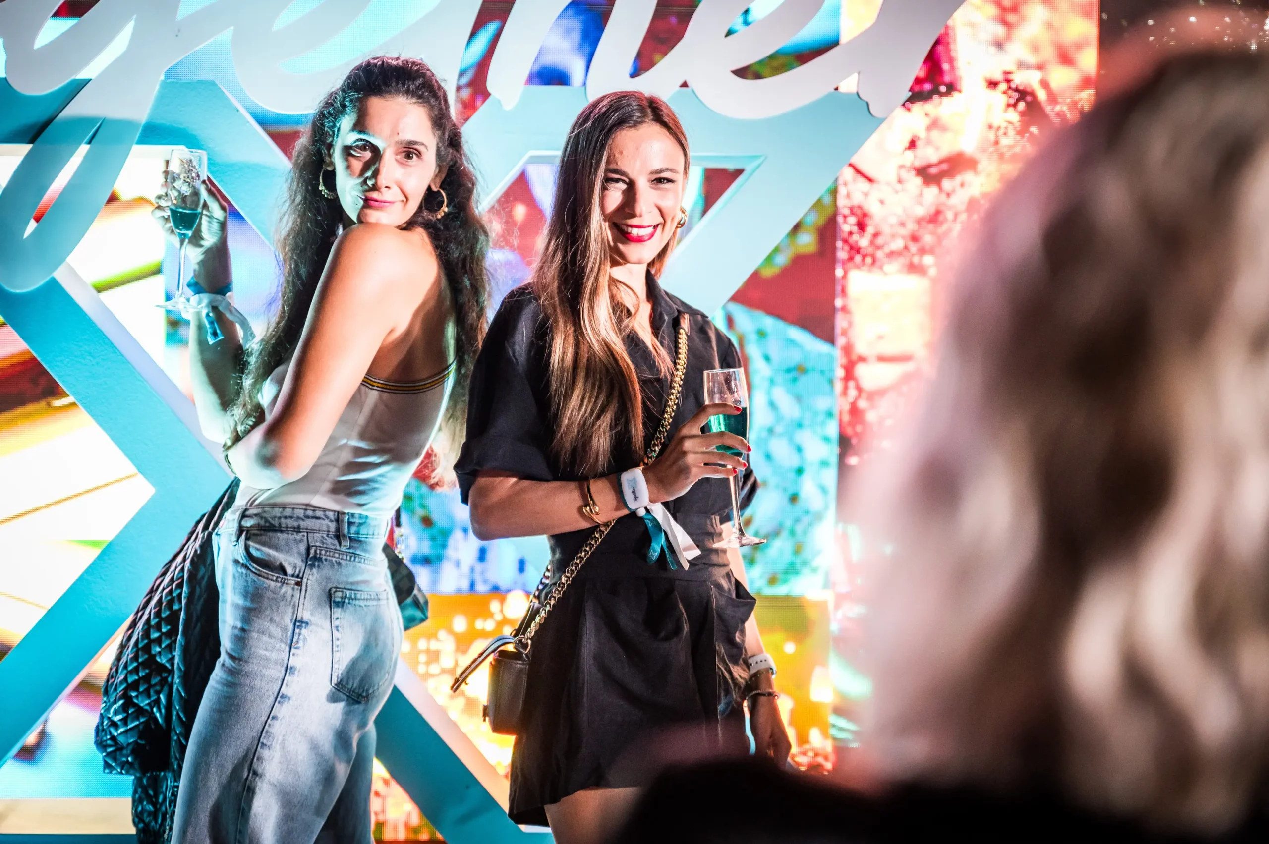 Two women posing with champagne glasses at a vibrant event backdrop. BattleRoyalStudios