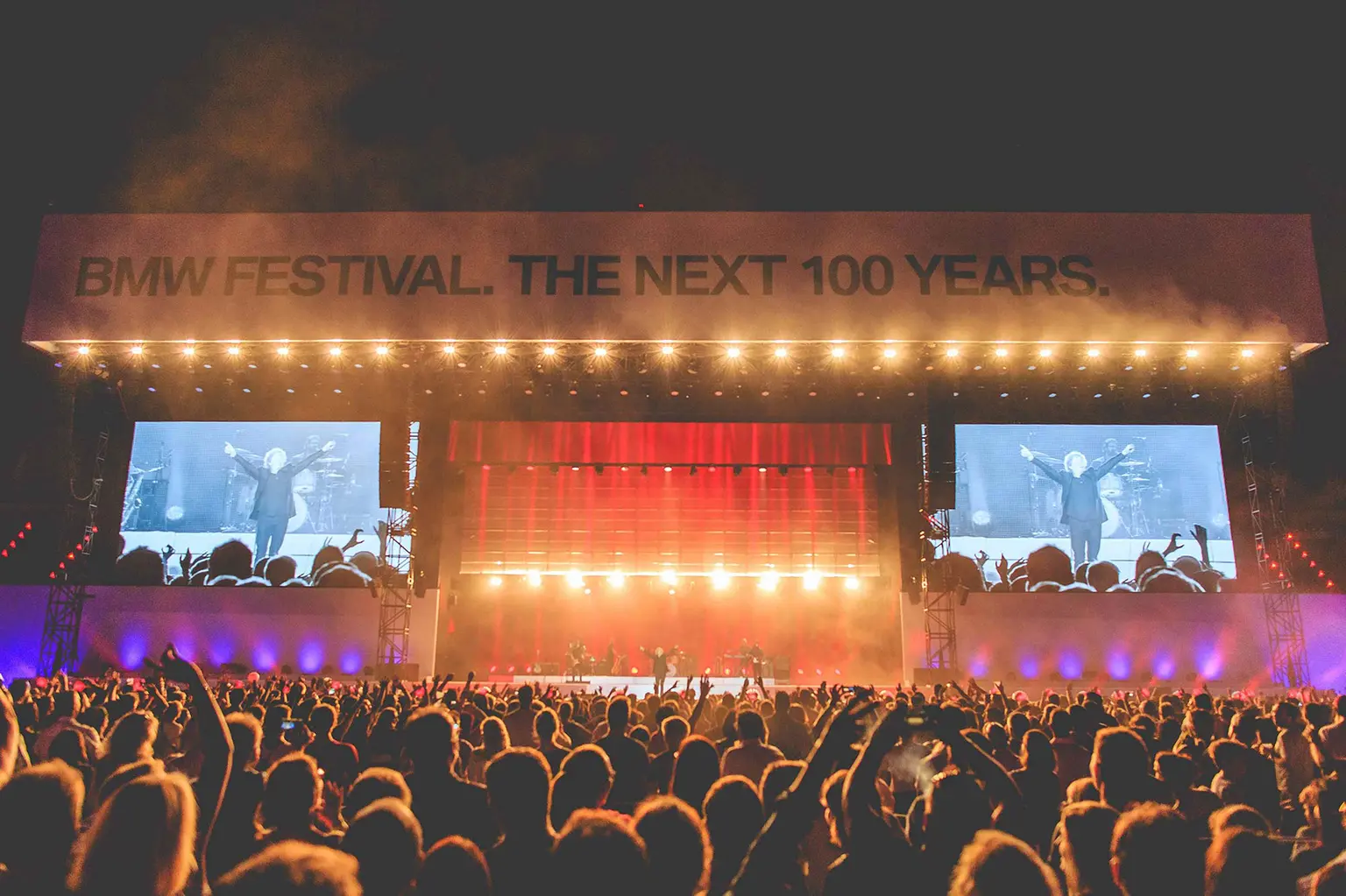 A large crowd attends a concert at the BMW Festival, with bright lights and large screens showing the performer on stage. The banner reads "BMW Festival. The Next 100 Years." Battle Royal Studios