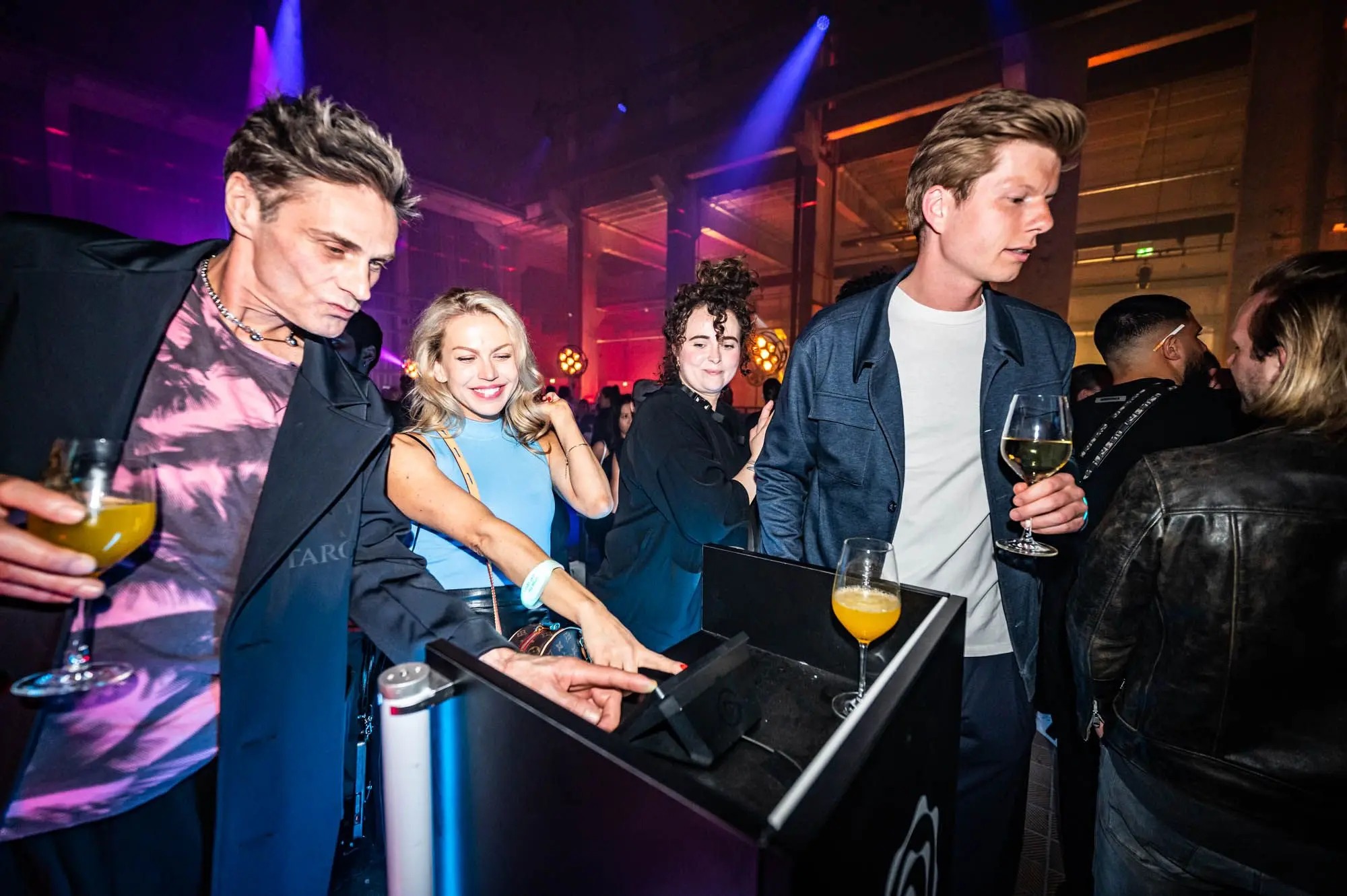 Guests with drinks gather around an interactive table in a dark venue with purple lights, one guest touching the table surface.