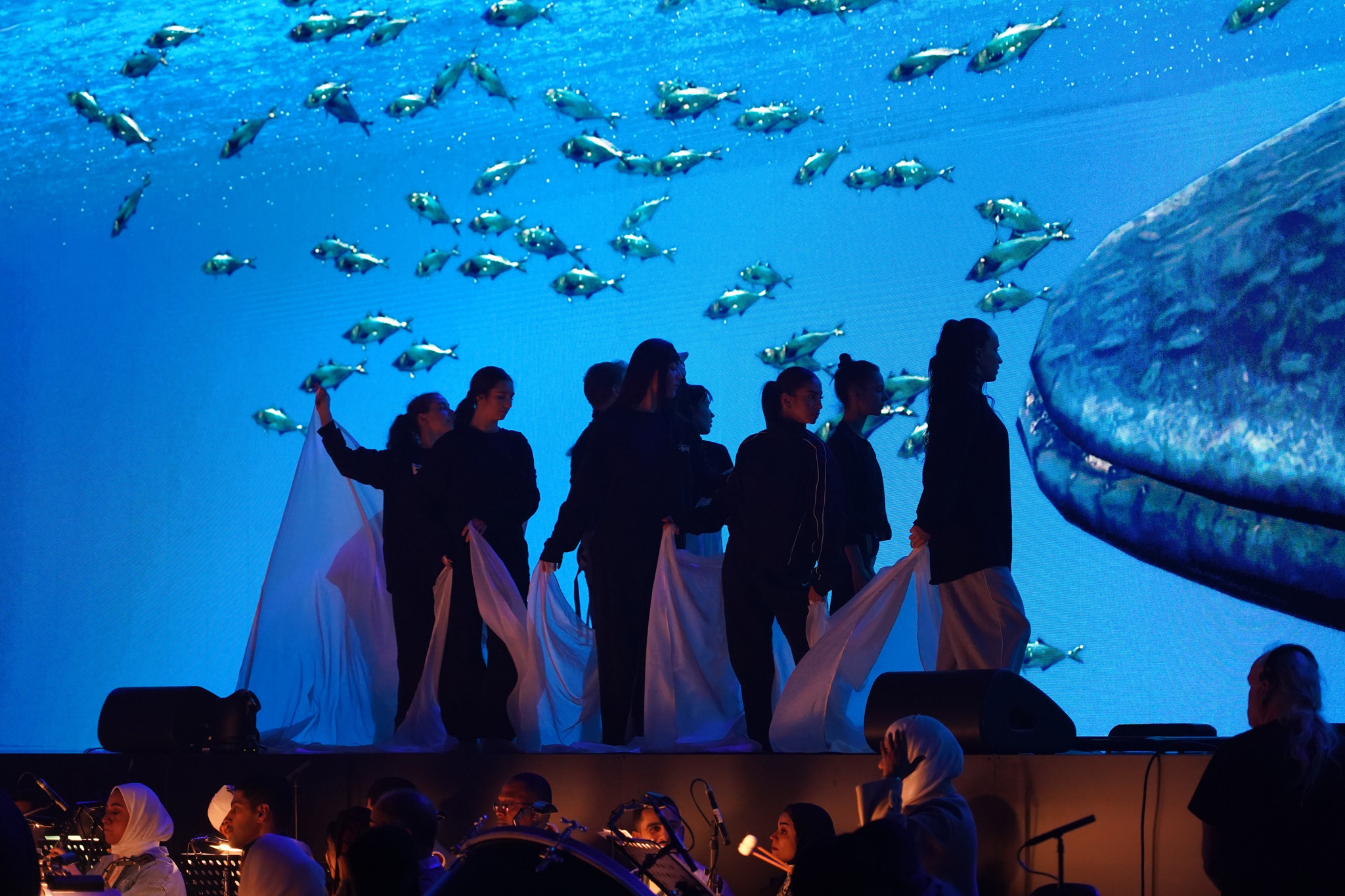 Dancers in dark clothing hold flowing white fabric on stage in front of an underwater projection of fish and a whale, with musicians in the foreground.