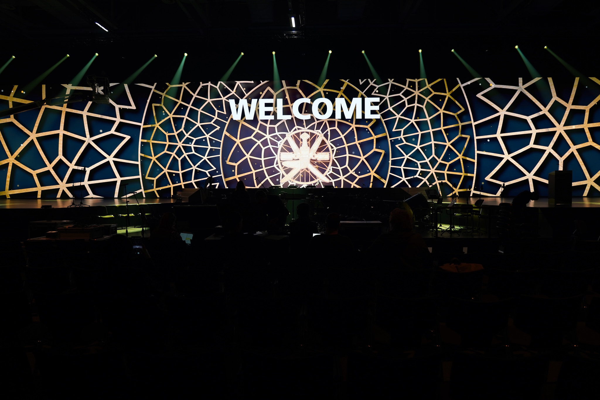 A long LED wall with gold geometric patterns and the word WELCOME stands behind a stage, with green light beams above and a dark hall in front.