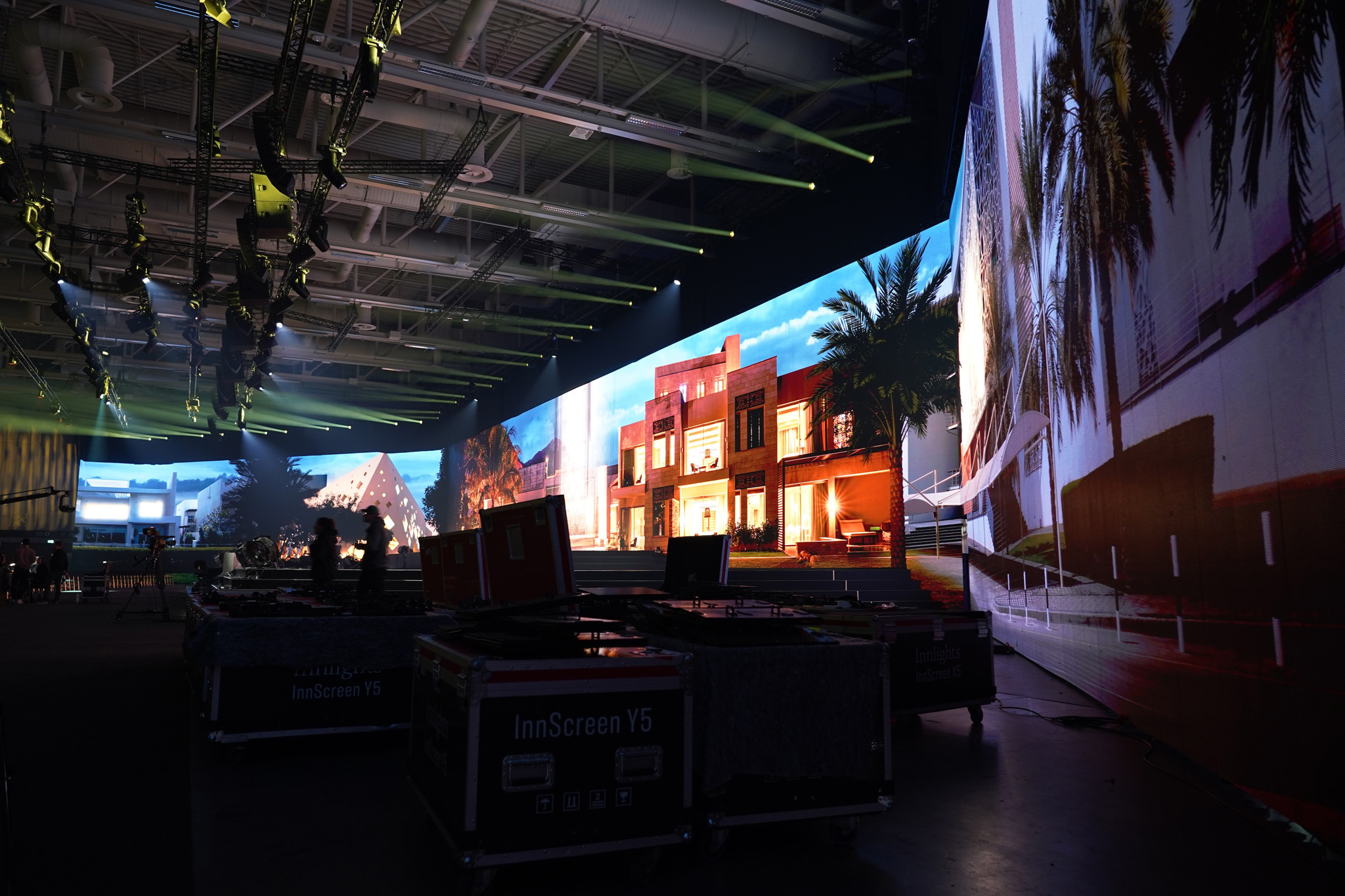 An LED wall shows a town with palm trees and low buildings along one side of a dark exhibition hall, with equipment cases and lighting rigs.