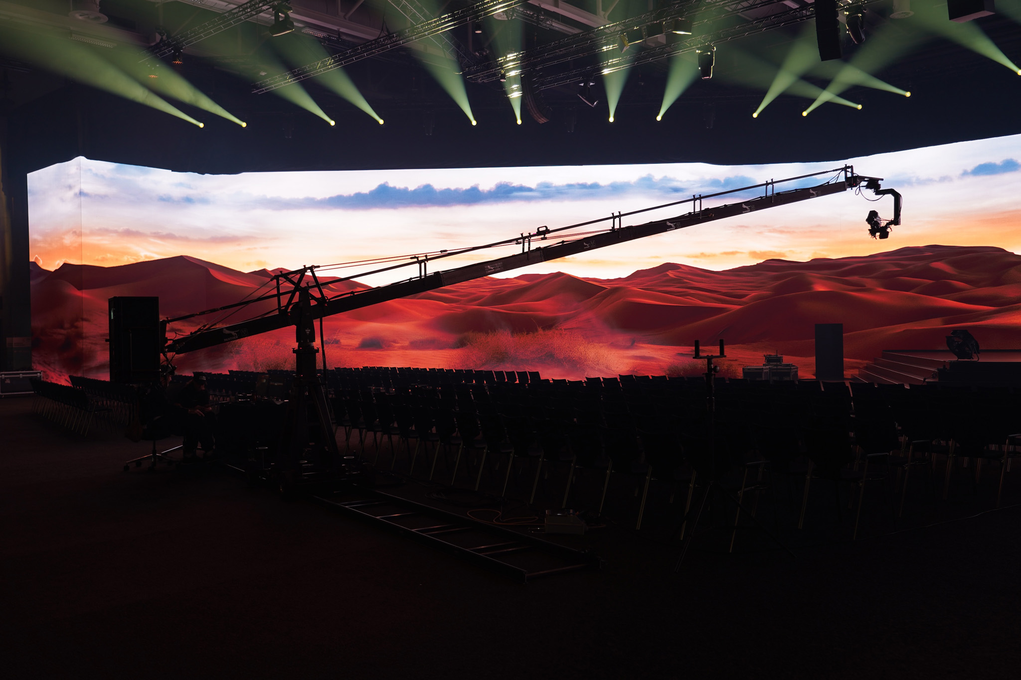 A camera crane extends across a dark hall towards a curved LED wall showing red desert dunes at sunset, with green light beams above.