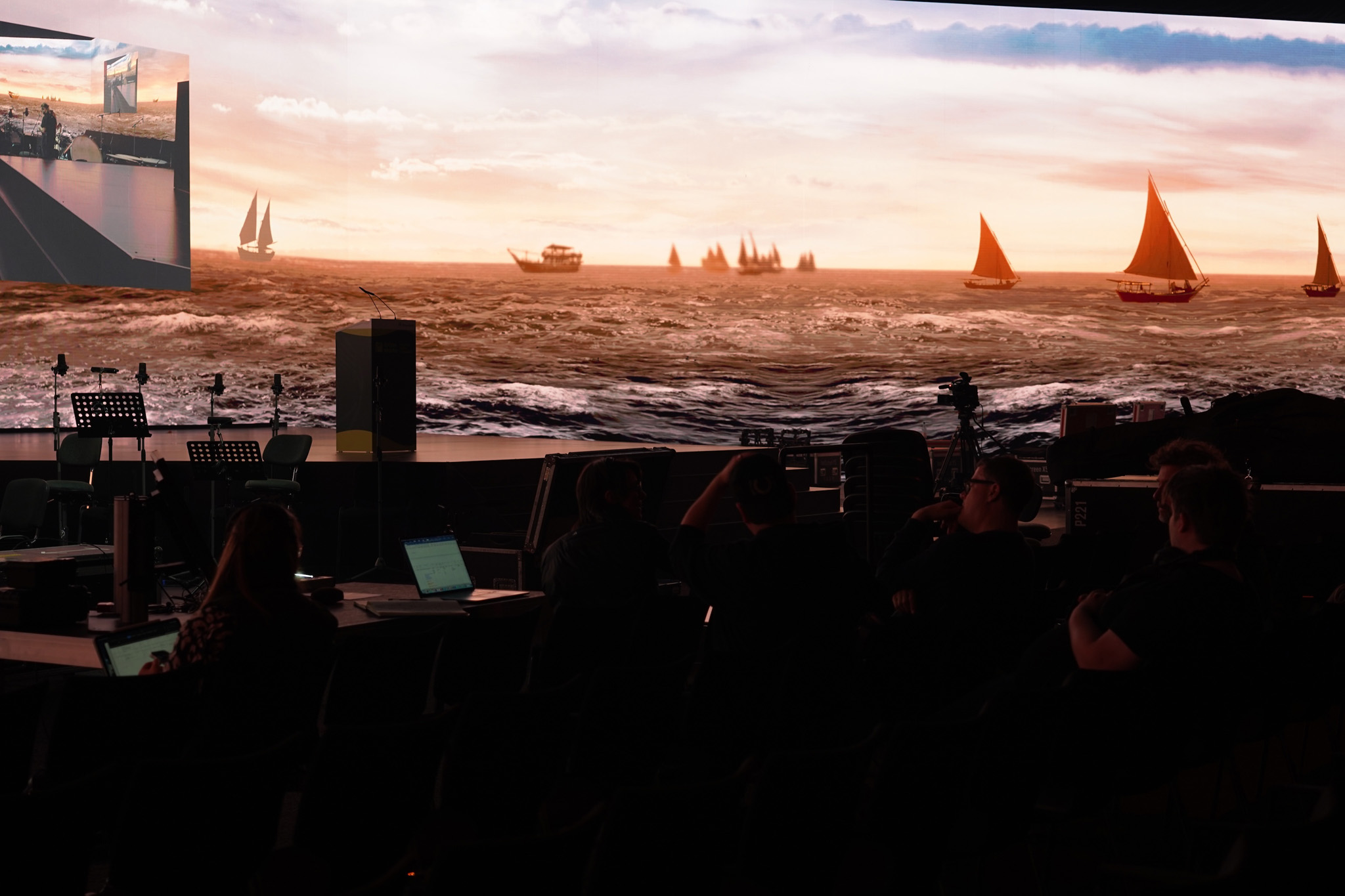 A panoramic LED wall shows sailing boats on a sunlit sea above a dark stage where musicians and crew are seated.