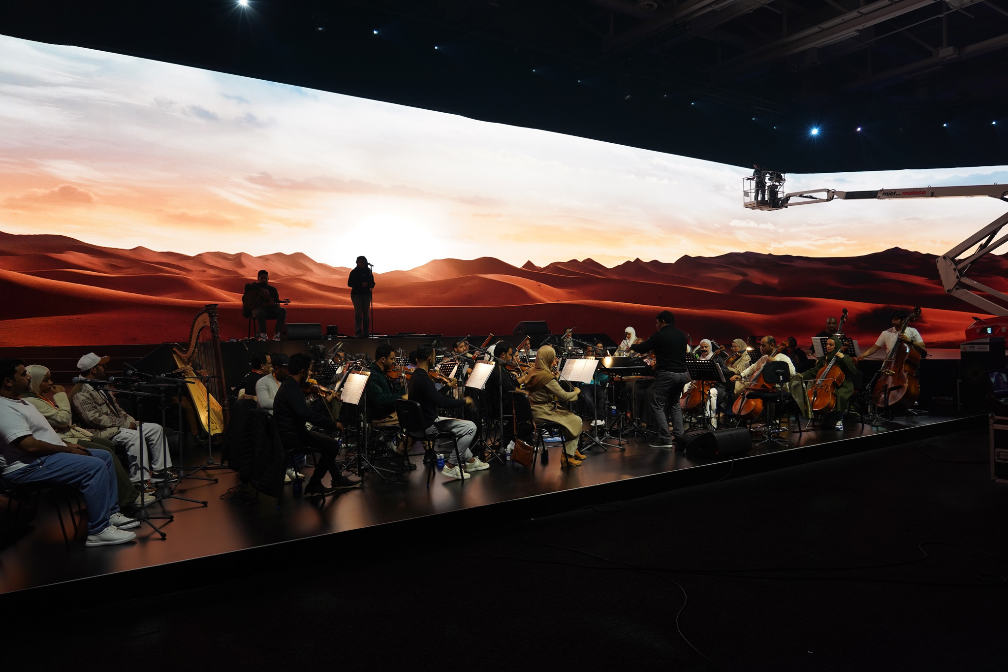 An orchestra sits on a long stage in front of a curved LED wall showing red desert dunes under a pale sky, with a camera crane above.