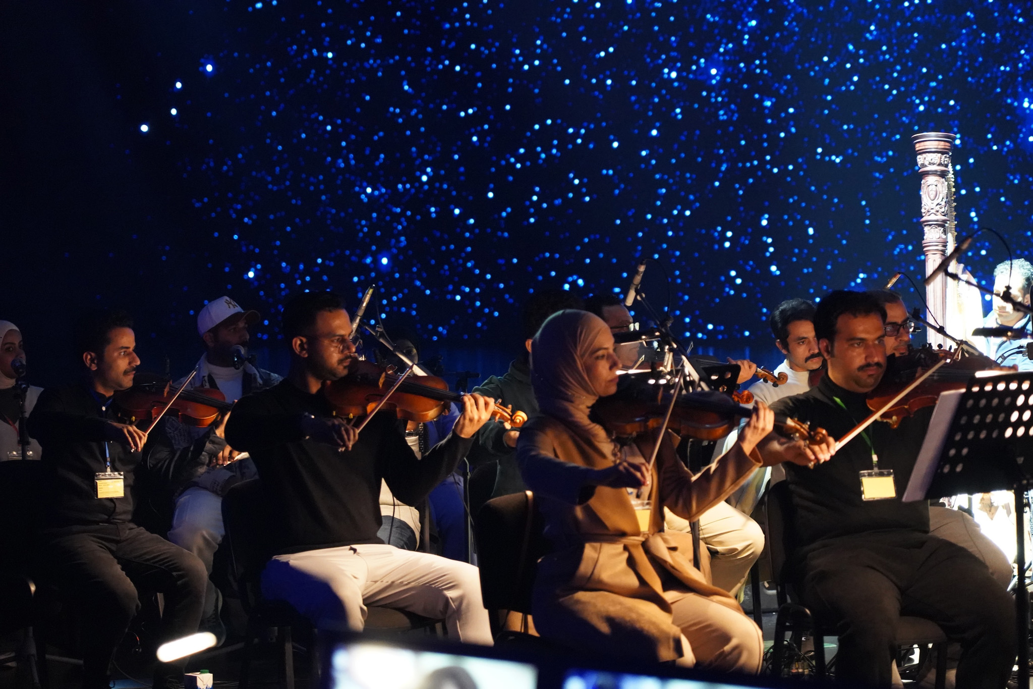 A string section plays violins on stage under blue light, with a starry sky on the LED wall behind them.