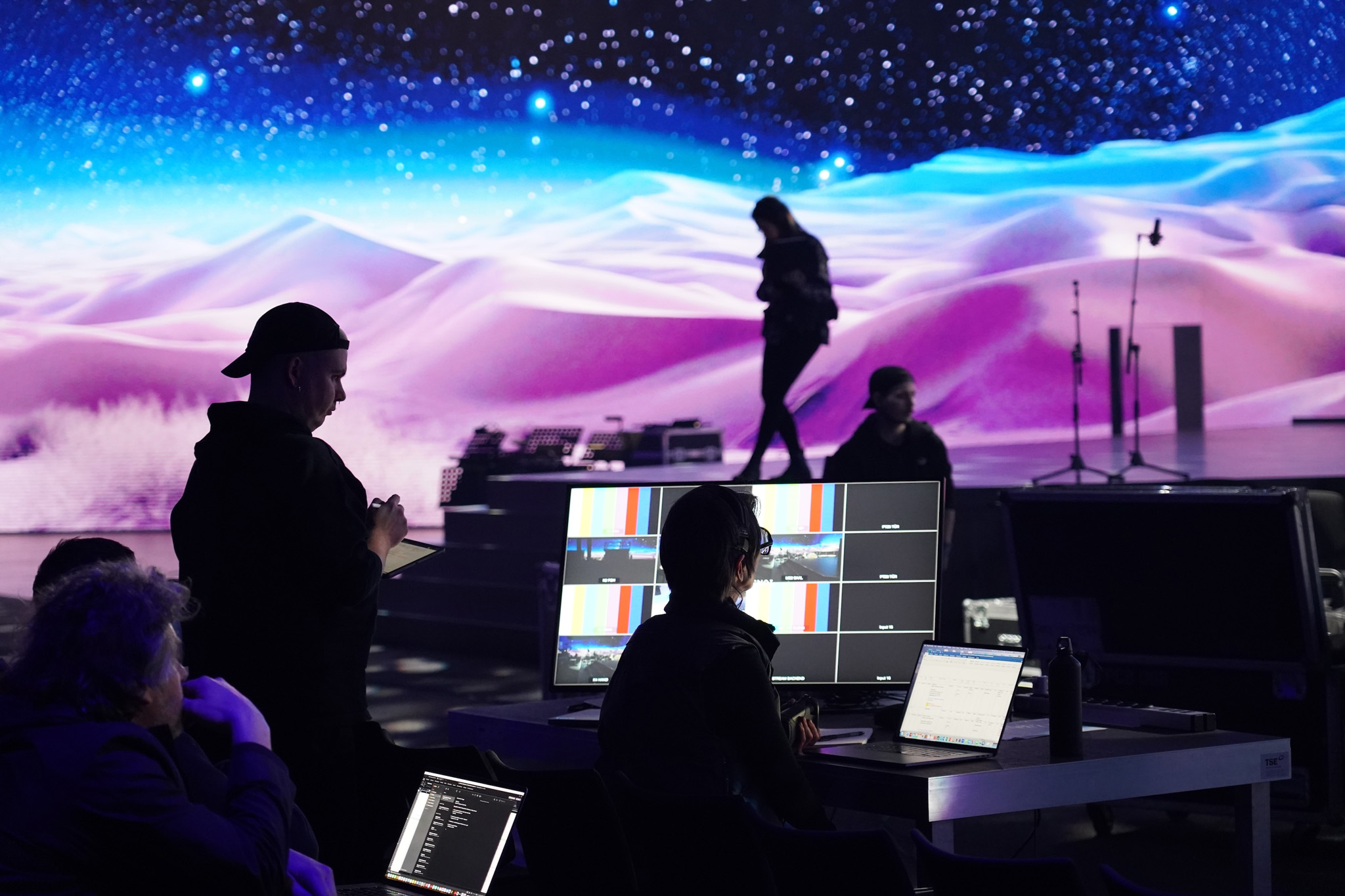 Technicians work at a control desk with monitors and laptops in front of an LED wall showing pink desert dunes under a starry sky, with a person standing on stage.