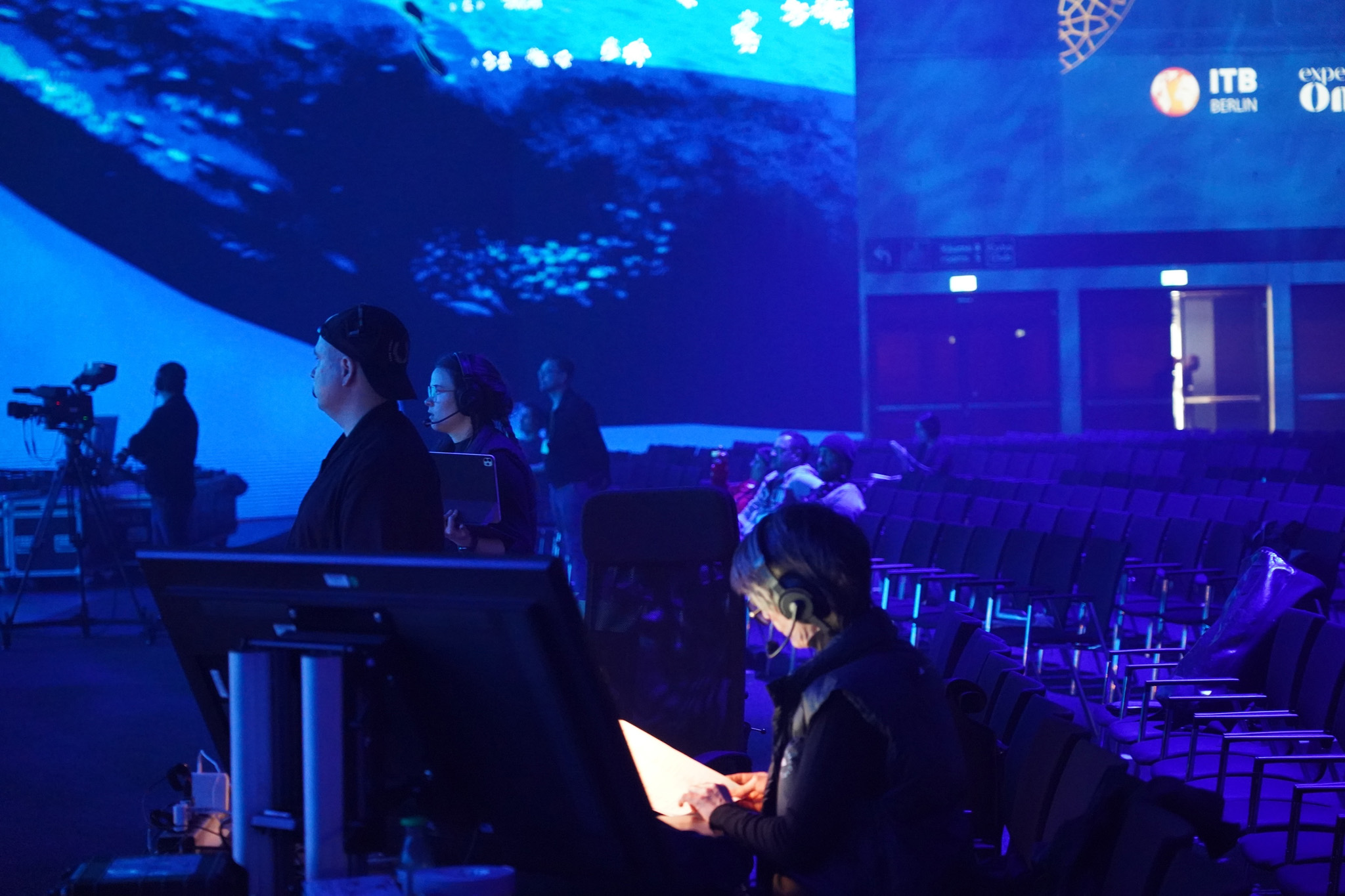 Crew members wearing headsets work at a desk in front of a blue underwater scene on the LED wall, with a camera operator and empty seats nearby.