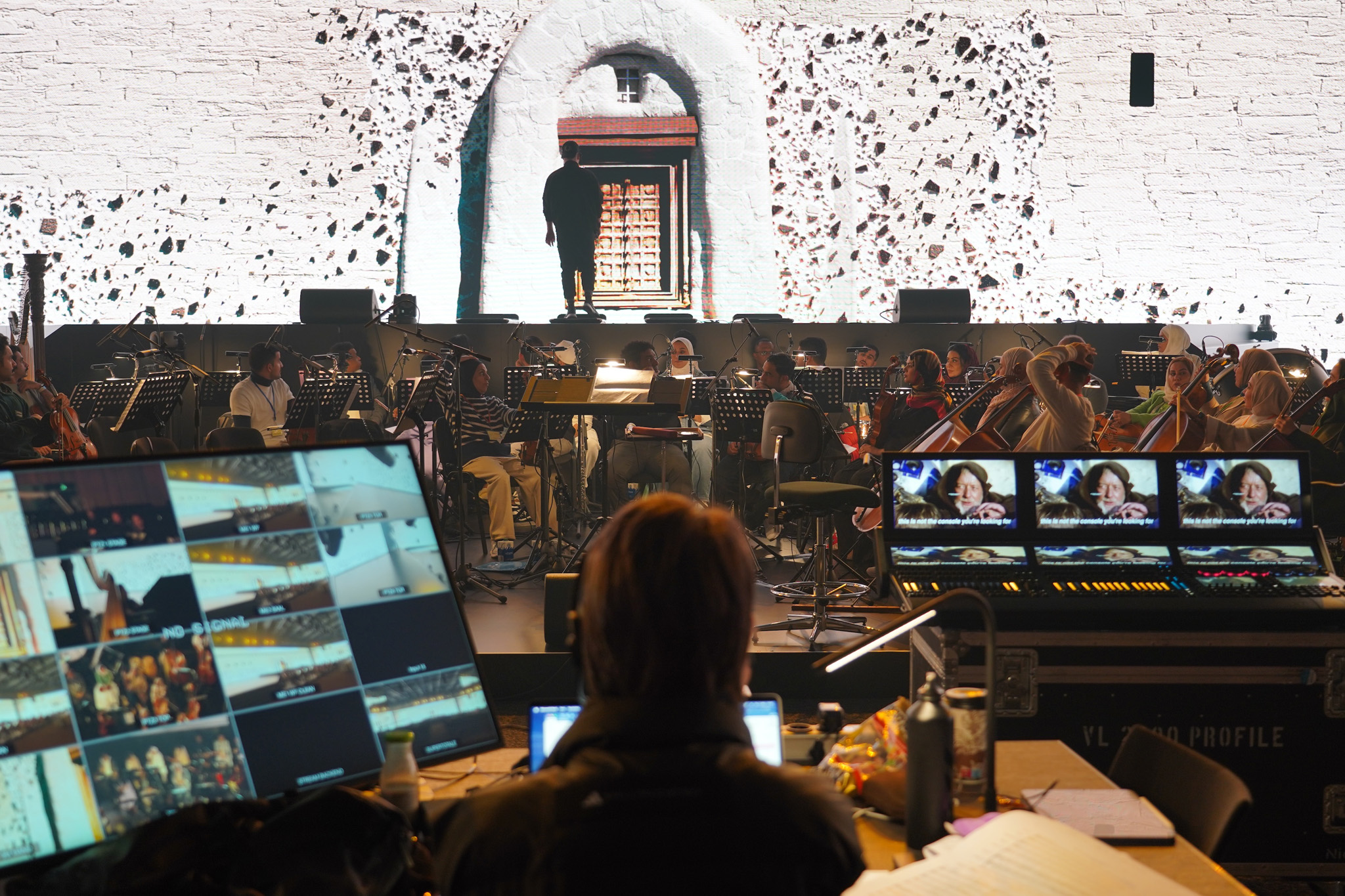 A technician sits at a desk with video monitors and a mixing console, facing a stage where an orchestra rehearses in front of a white wall with an arched doorway.