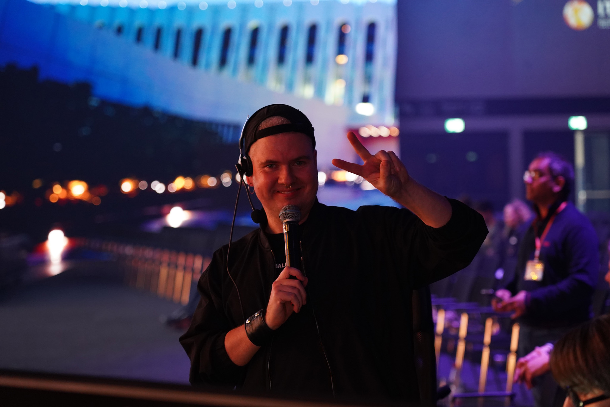 A crew member wearing a headset and cap speaks into a microphone and gestures with one hand, with lit stage graphics and other crew behind him.