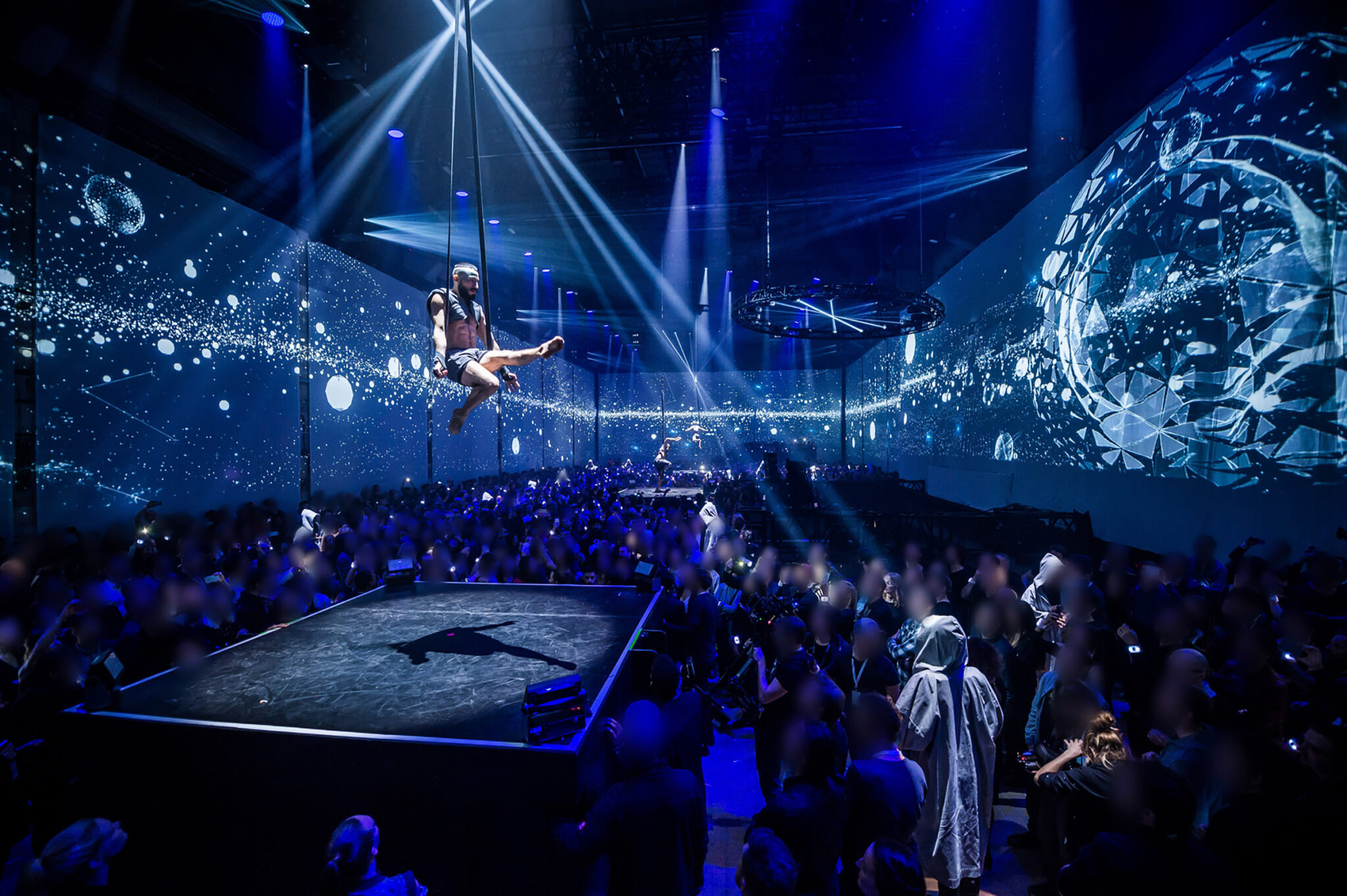 An acrobat hangs from straps high above a dark stage, legs extended, while projections of glowing particles and a large sphere cover the surrounding walls and a crowd watches from below.