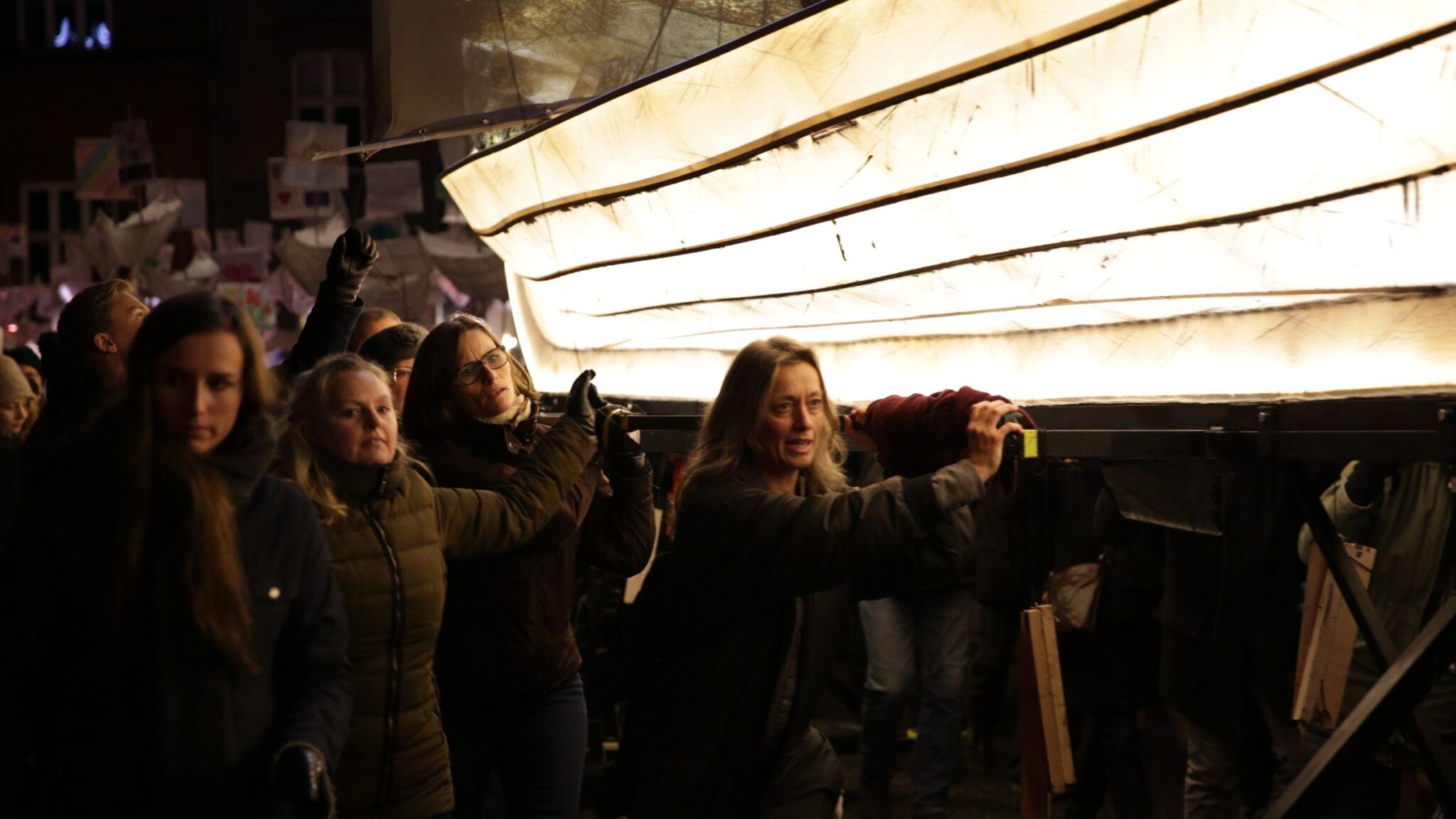 People carrying a large illuminated structure resembling a boat during a nighttime event. Battle Royal Studios.