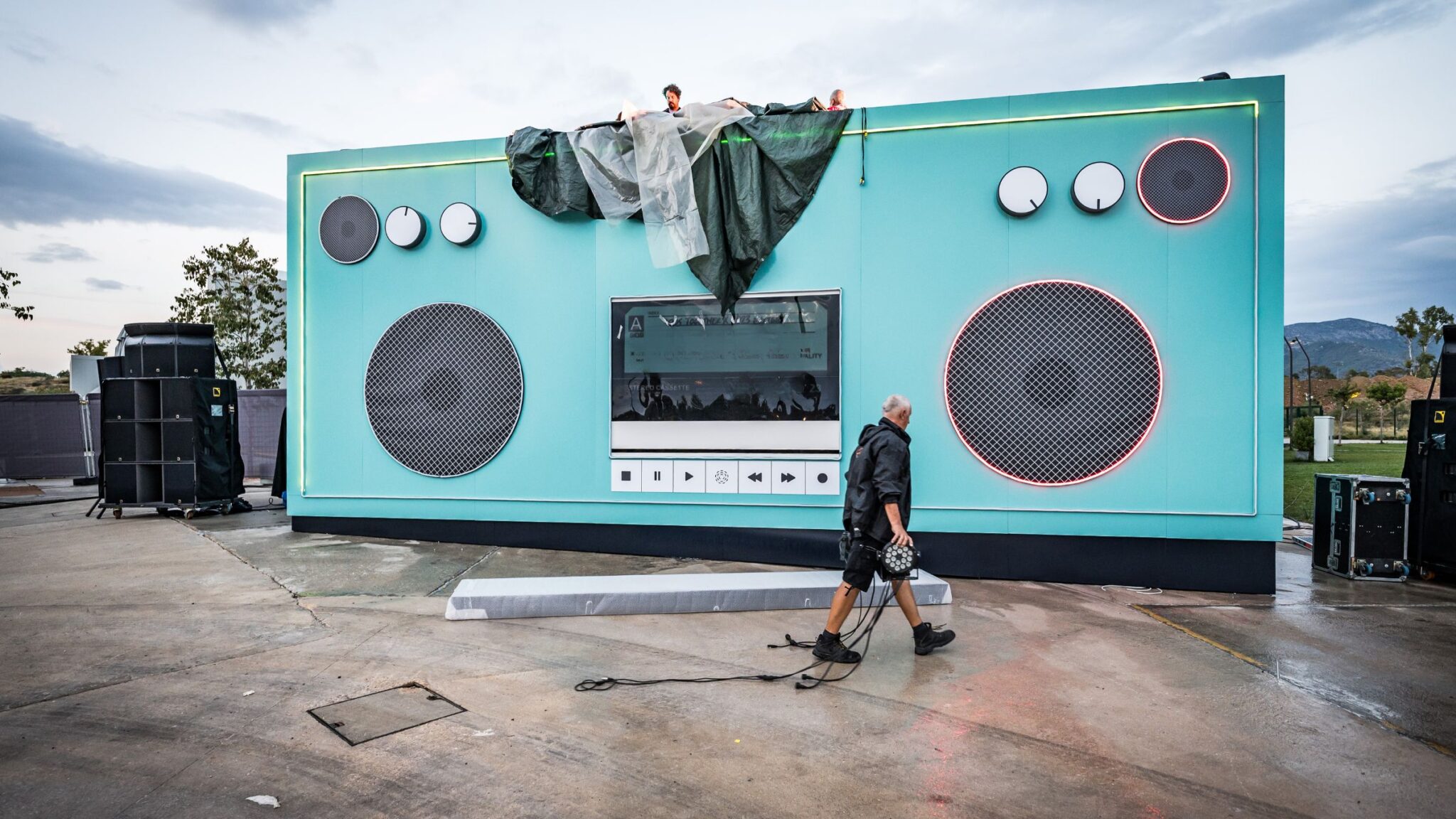 A large stage resembling a boombox is being set up, with workers adjusting equipment and covering parts of the structure. Battle Royal Studios