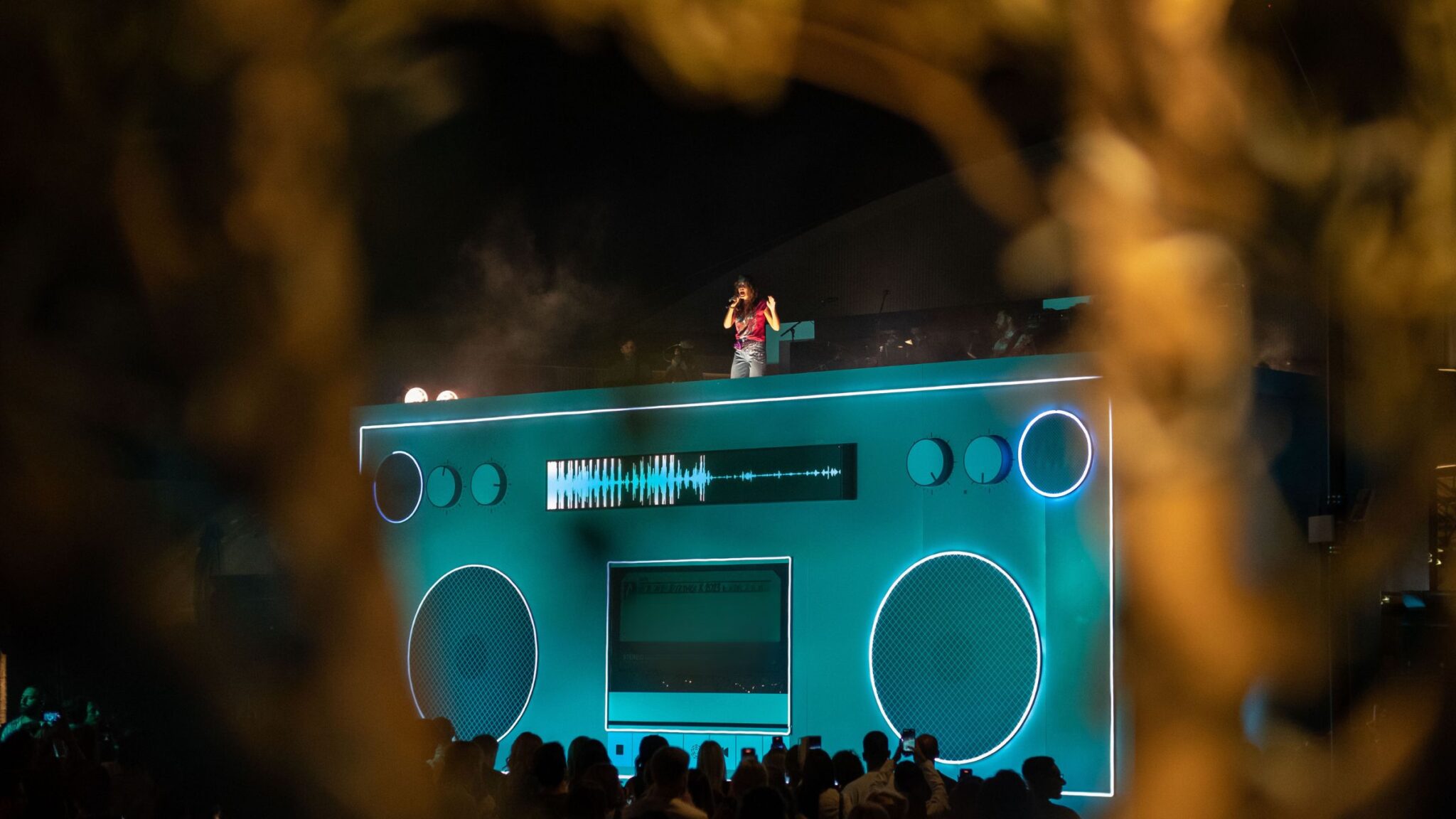 A performer sings on top of a stage designed as a large boombox, with glowing neon details, while a crowd watches below. Battle Royal Studios