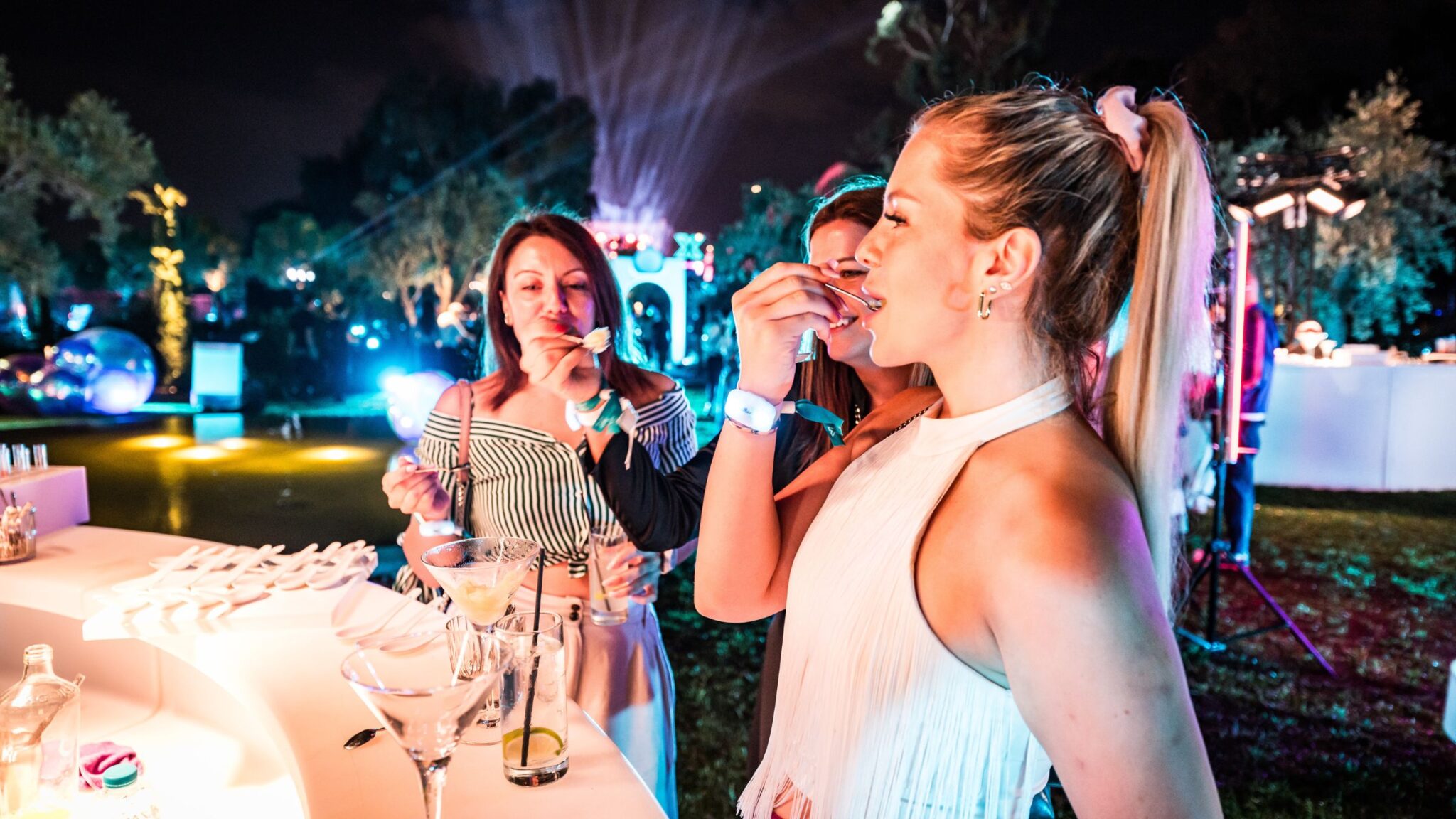 Three women enjoying food and drinks at an outdoor evening event, with colorful lights and a festive atmosphere in the background. Battle Royal Studios