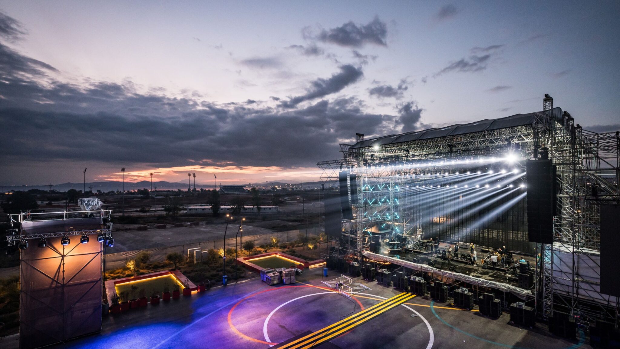 An outdoor stage is being set up with bright lights shining across the venue as the sun sets in the background. Battle Royal Studios