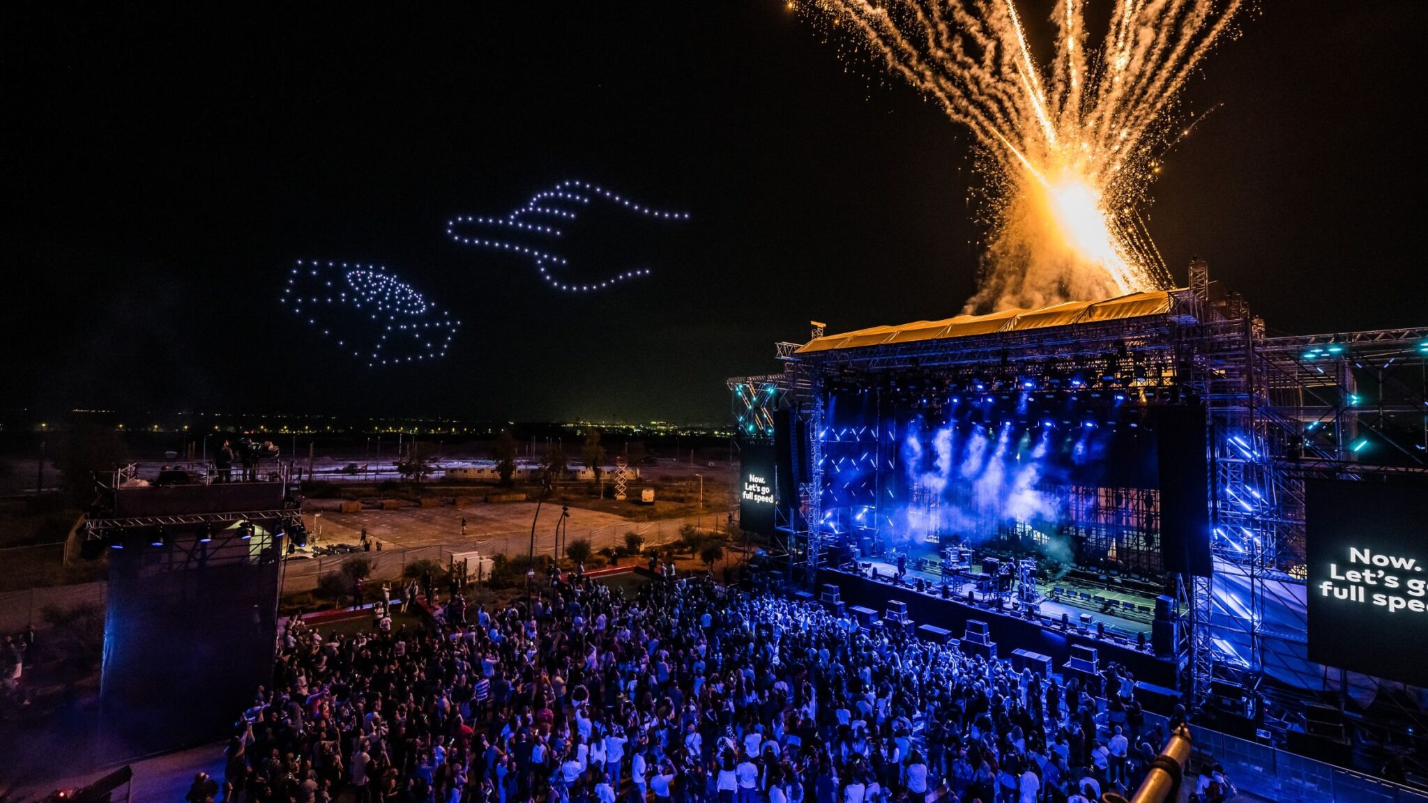 A large crowd watches a nighttime performance on an outdoor stage, with fireworks and a drone light display forming shapes in the sky. Battle Royal Studios