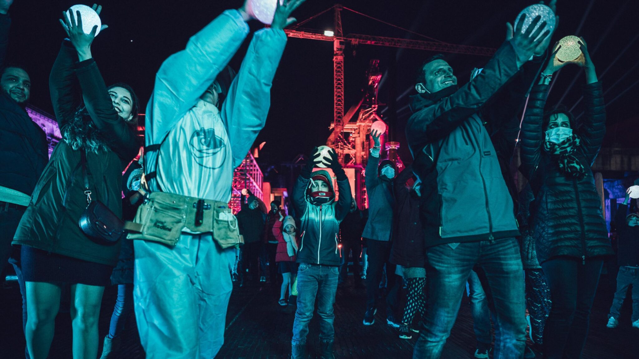 People raising glowing spheres above their heads during an outdoor nighttime event, with colorful lights and cranes in the background. Battle Royal Studios.