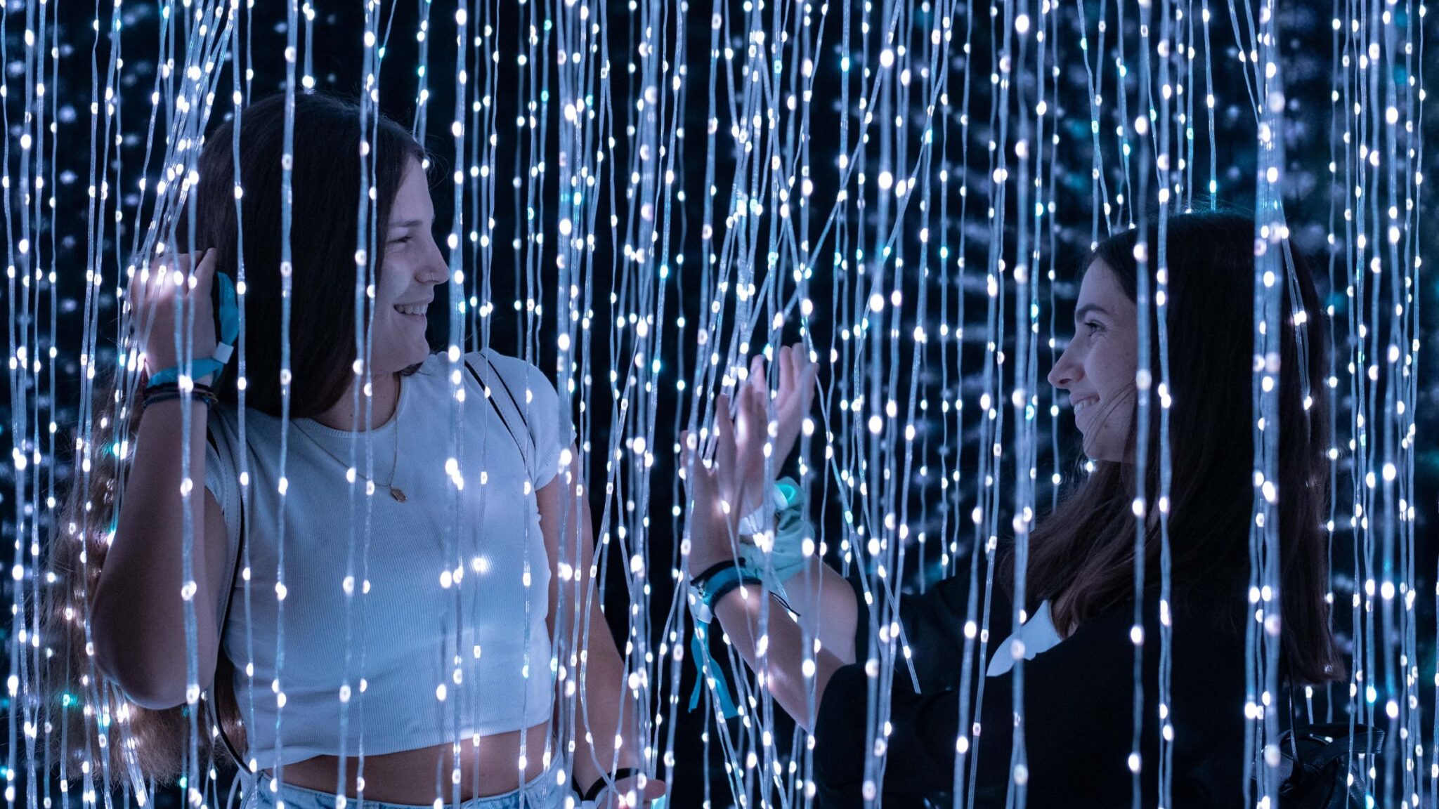 Two women smiling at each other while standing among hanging strings of glowing lights. Battle Royal Studios.