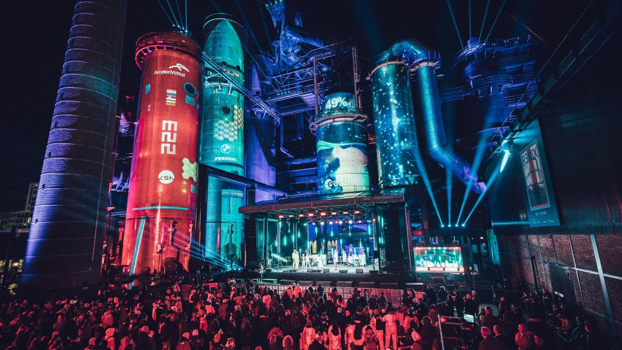 Crowd watching a performance at night, with large illuminated industrial silos and beams of light creating a vibrant atmosphere. Battle Royal Studios.