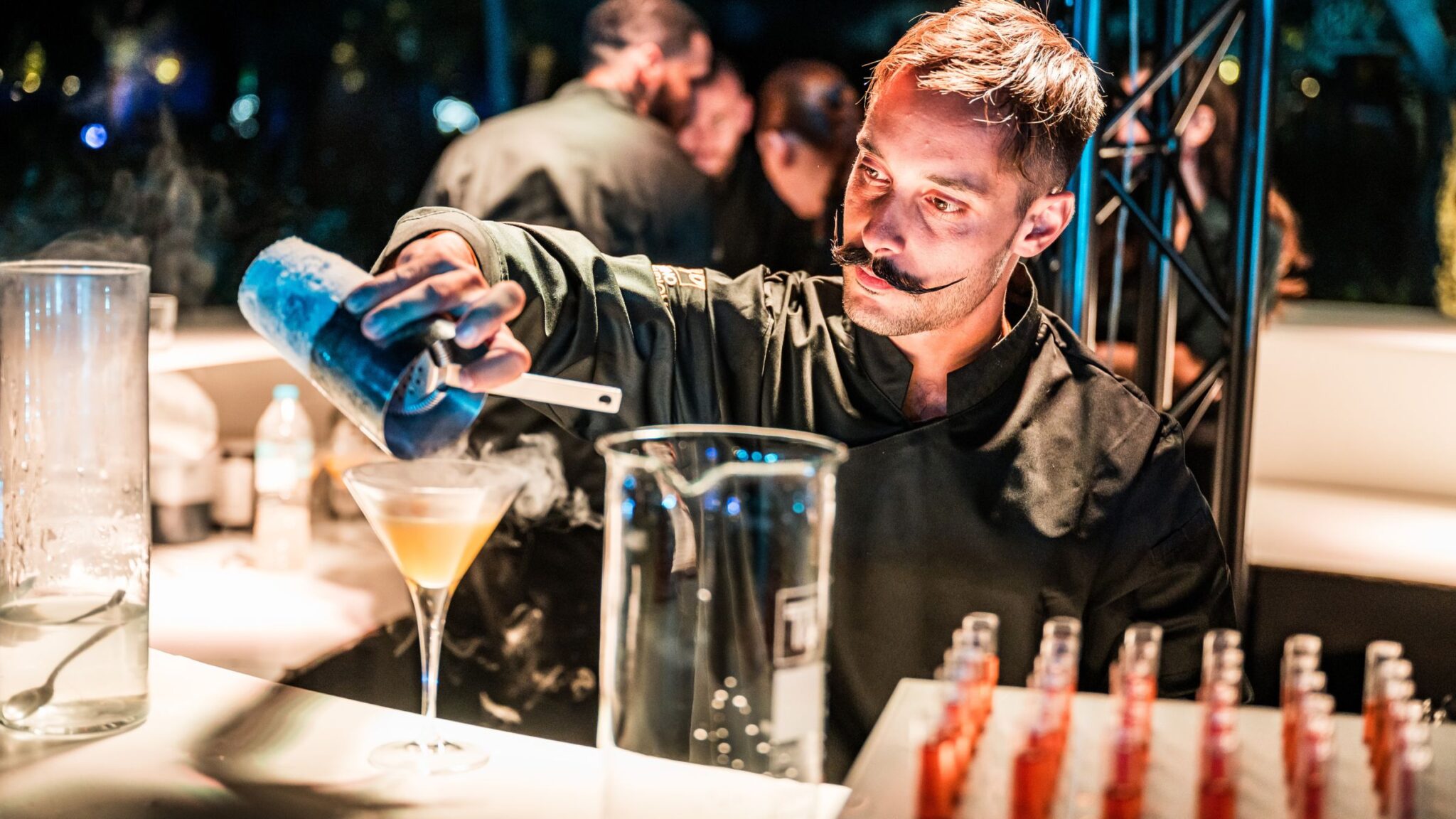 A bartender with a styled mustache carefully pouring a drink into a cocktail glass at an upscale event, with smoking liquid nitrogen adding a dramatic effect. Battle Royal Studios