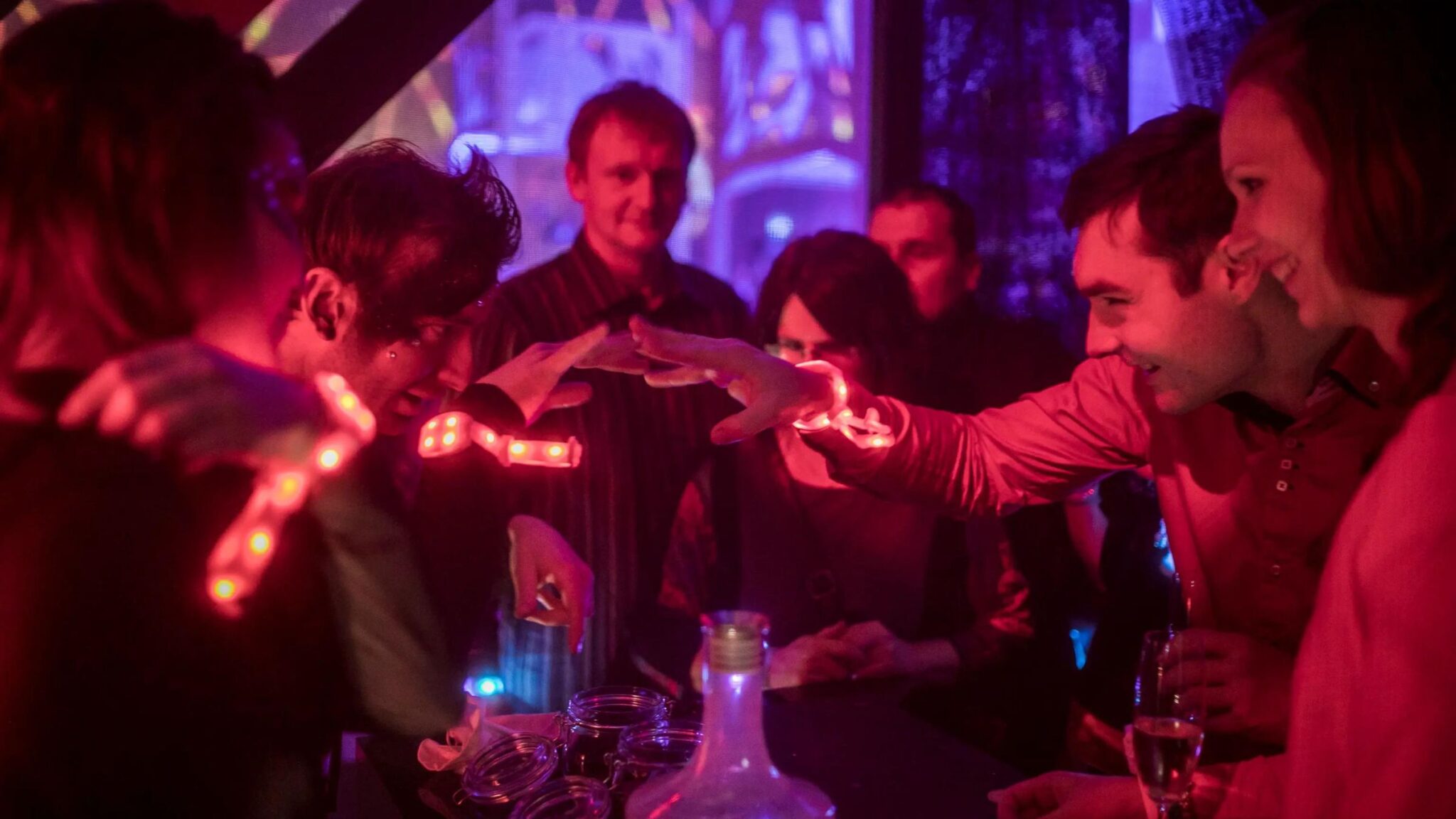 A group of people interacting and smiling at a party, illuminated by glowing red wristbands, in a colorful and vibrant environment. Battle Royal Studios