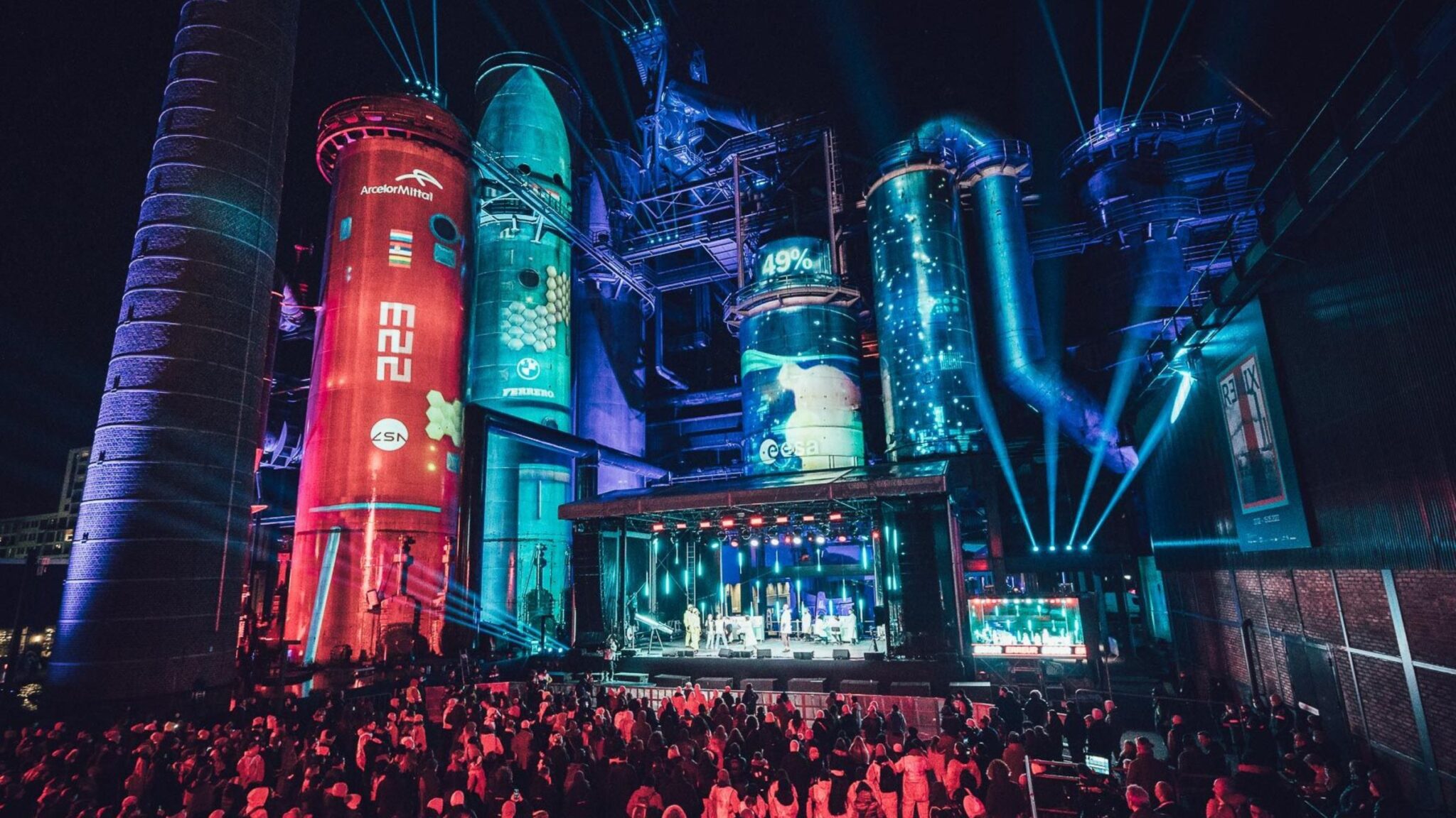 Crowd watching a nighttime performance in front of industrial silos illuminated with colorful projections and light beams. Battle Royal Studios.