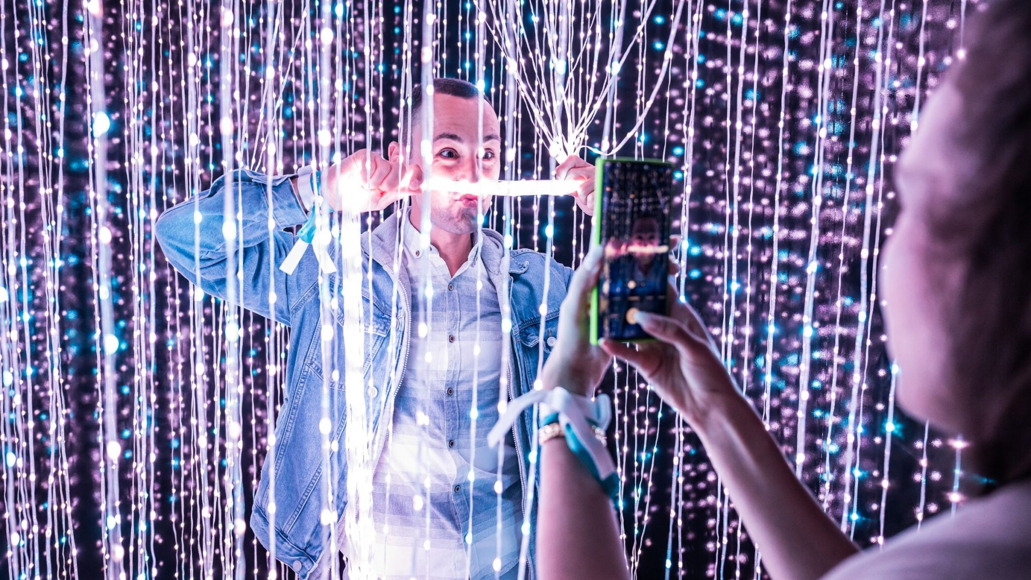 A man playfully holds a glowing light between his teeth, surrounded by strings of small lights, while someone takes his photo. Battle Royal Studios