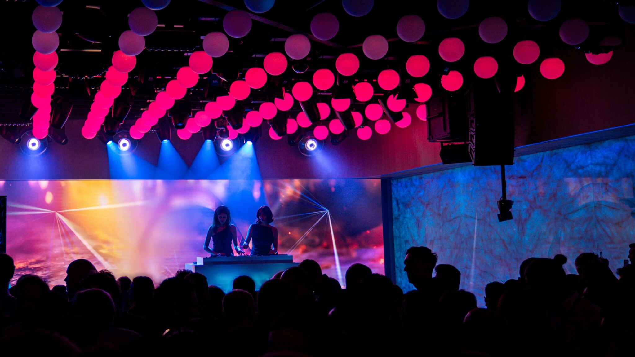 A DJ performance is taking place under rows of pink and red illuminated orbs, with a colorful backdrop and a crowd of people in the foreground. Battle Royal Studios