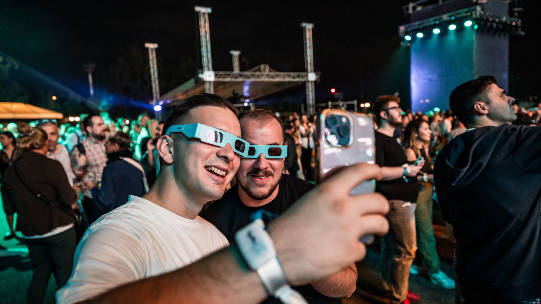 Two men wearing 3D glasses taking a selfie at an outdoor event, with a crowd in the background. Battle Royal Studios.