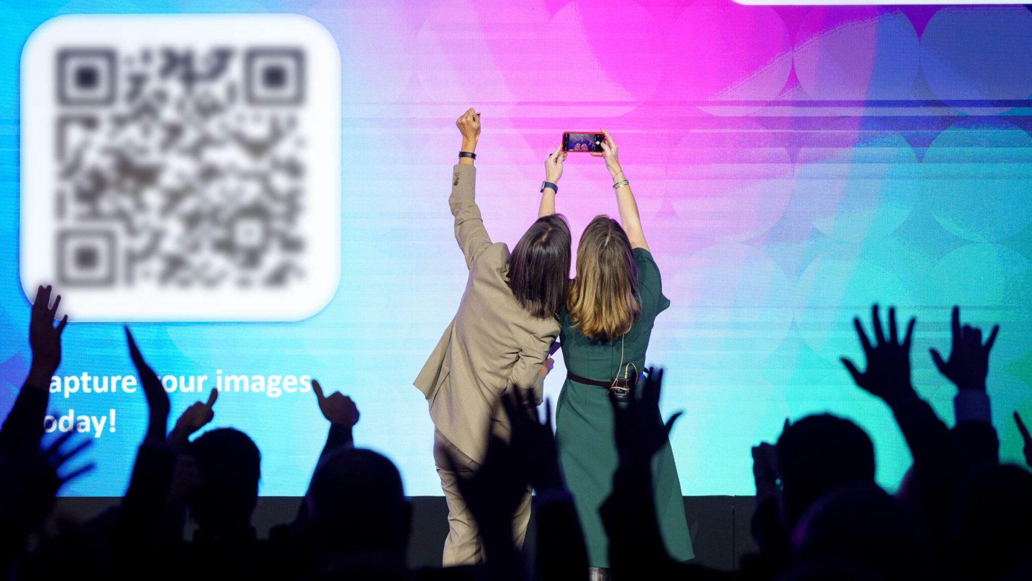 Two people take a selfie in front of a large screen displaying a QR code, with an audience raising their hands in the foreground. Battle Royal Studios