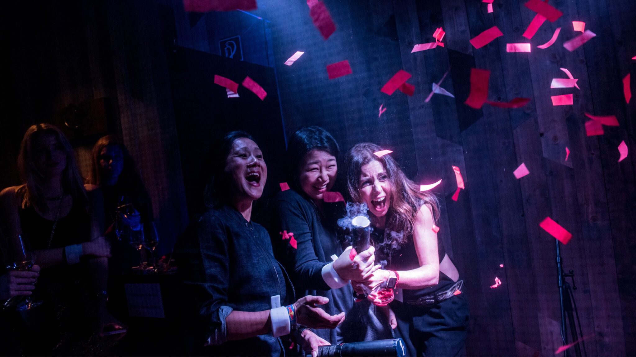 Three women joyfully celebrating at a party, with red confetti falling around them in a dark, vibrant setting. Battle Royal Studios