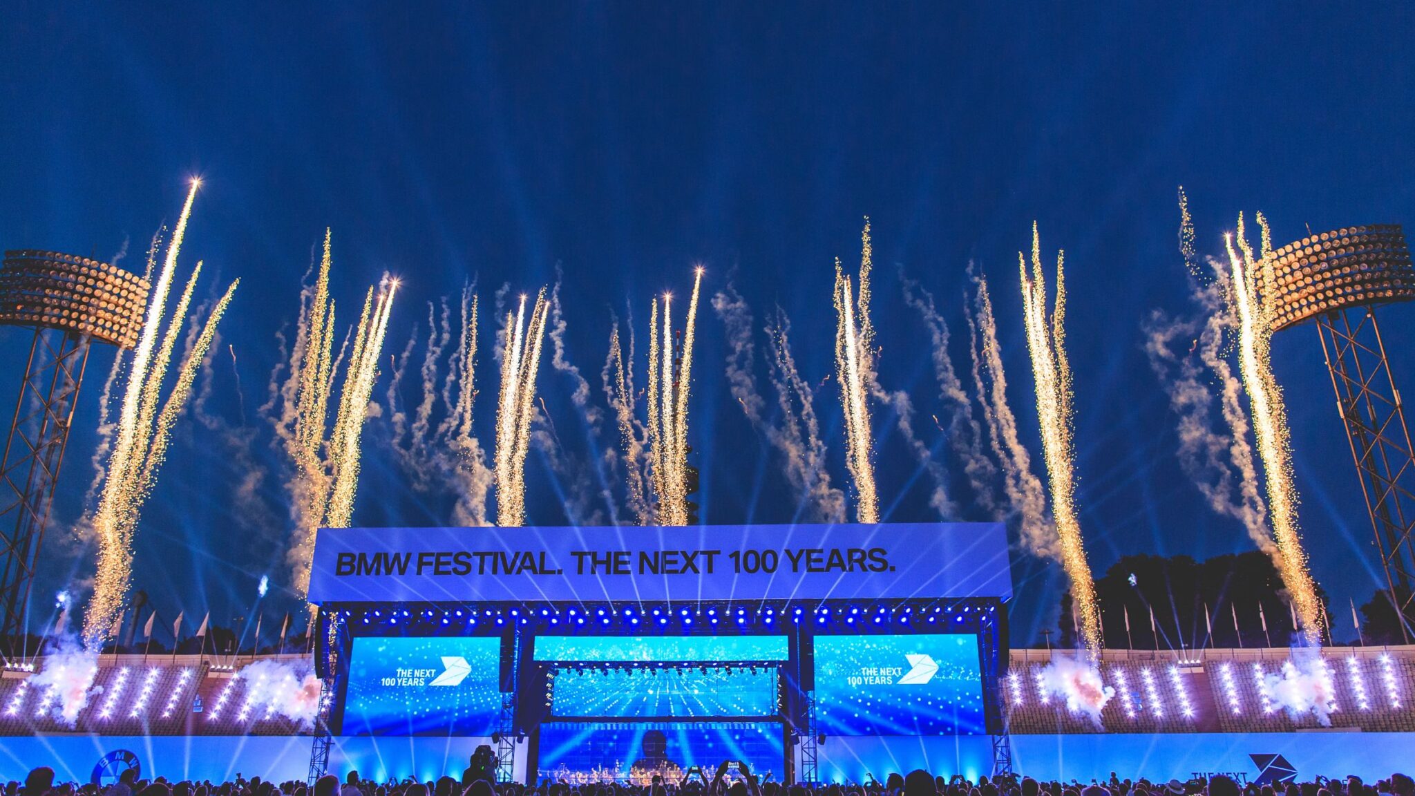 Fireworks light up the sky above the stage at the BMW Festival, with the sign "BMW Festival. The Next 100 Years." displayed prominently. Battle Royal Studios