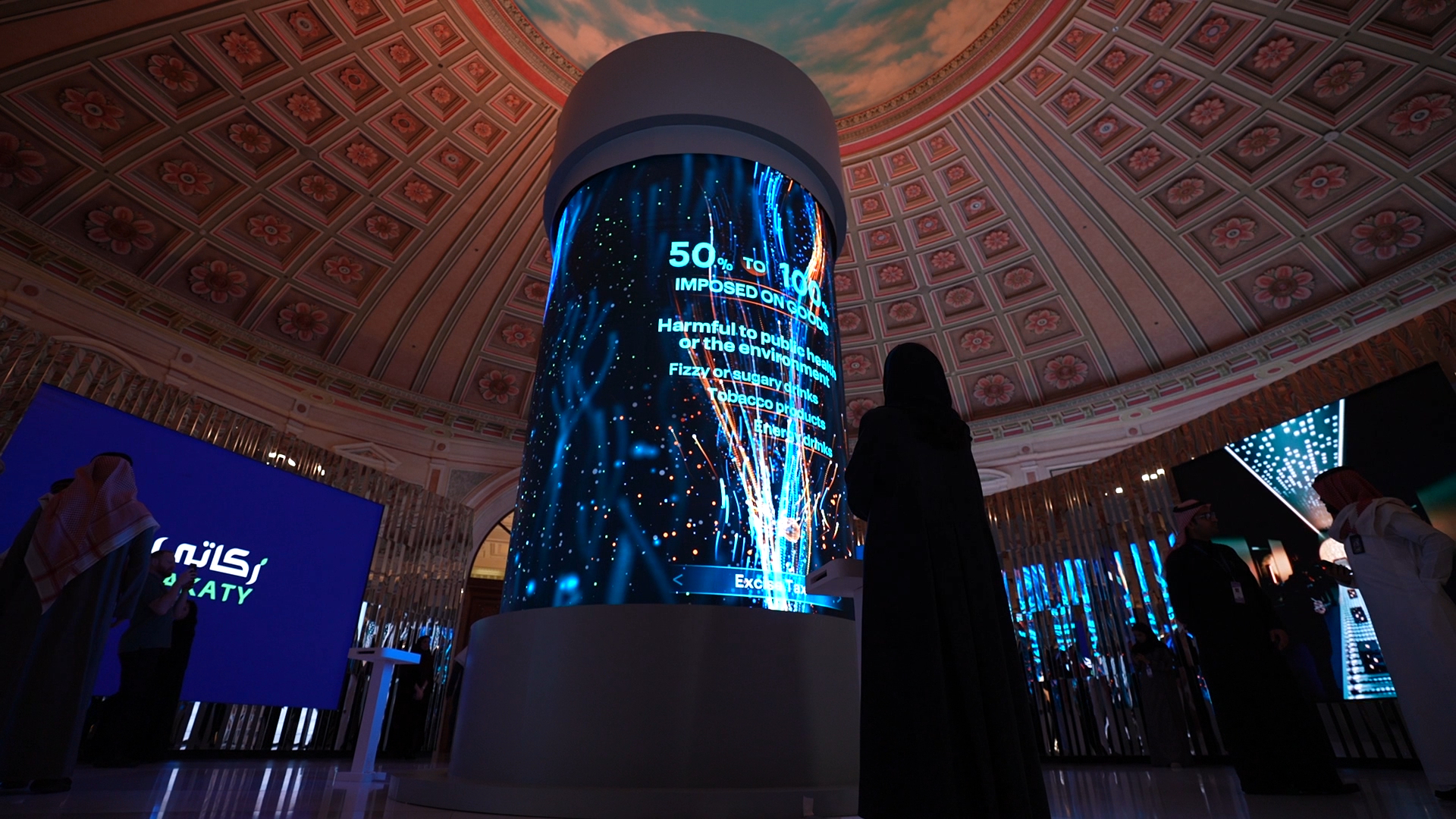 A person stands in front of a large cylindrical digital display showcasing animated graphics and text, set in an ornate hall with a decorative domed ceiling. Battle Royal Studios