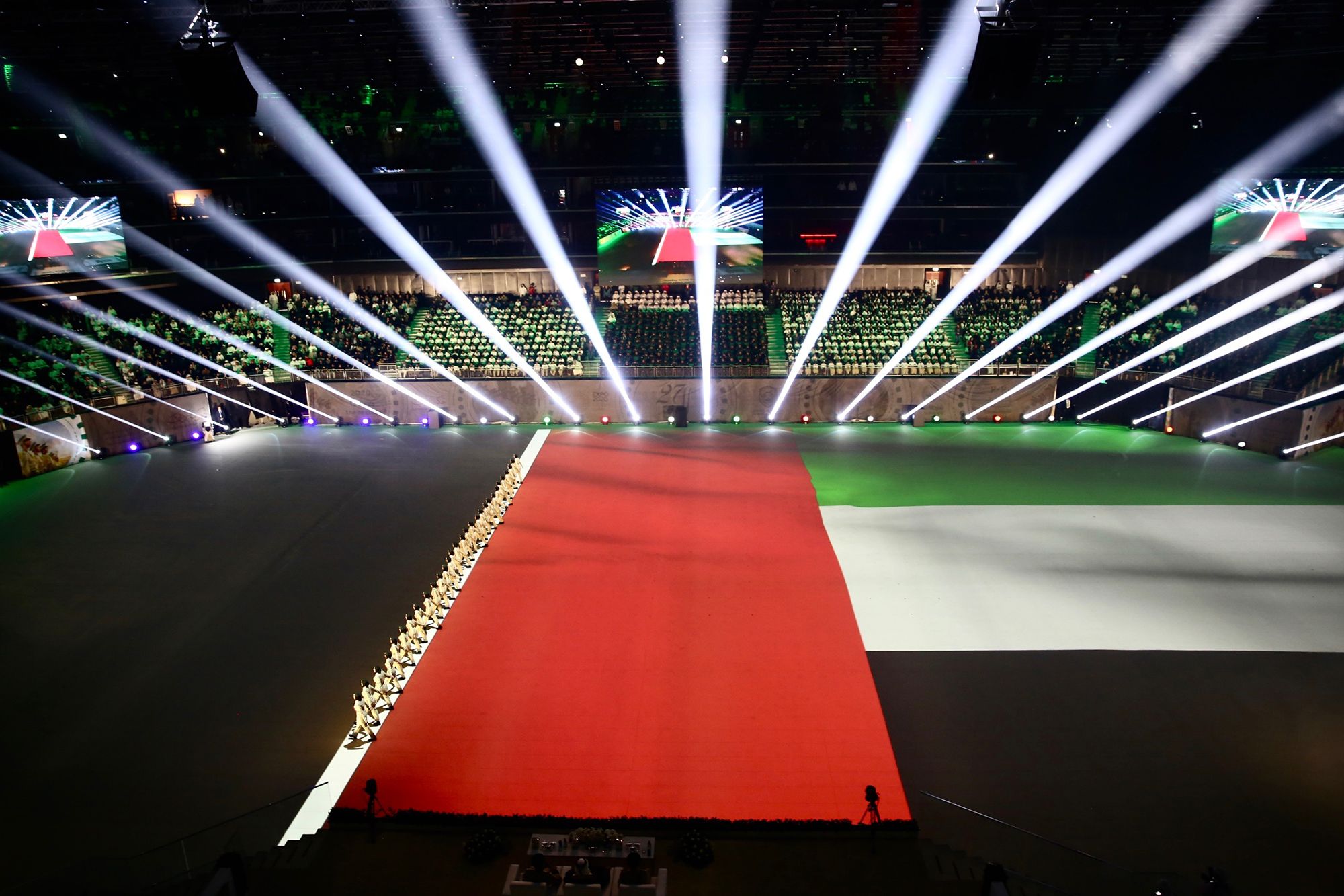 A grand indoor event featuring a massive UAE flag projection, bright spotlights, and an audience in the background. Battle Royal Studios