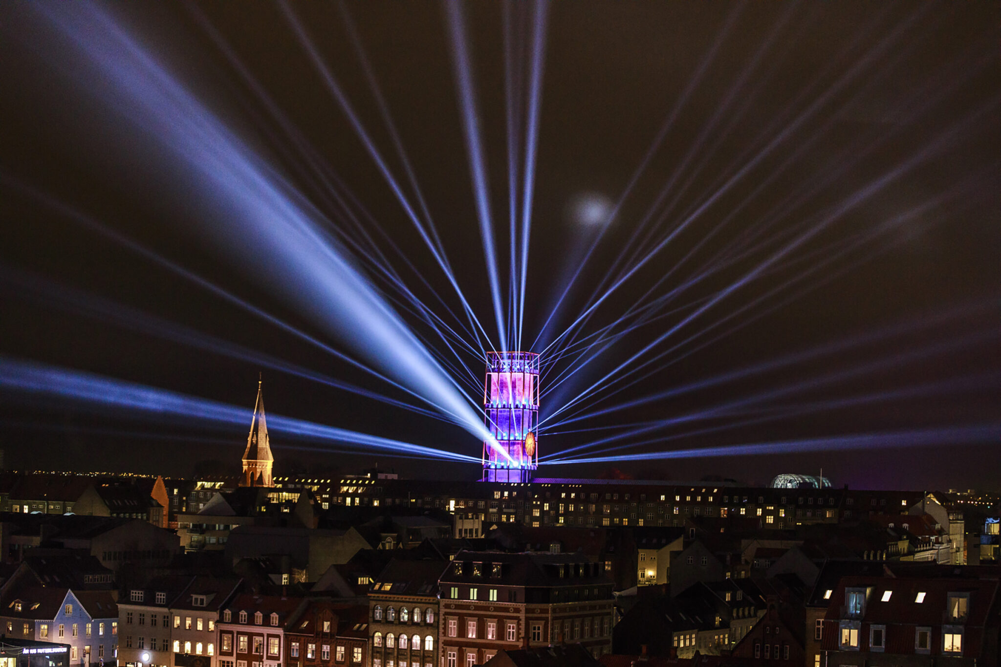 Beams of white light fan out from an illuminated tower structure into the night sky above a city of low rooftops and church spires.