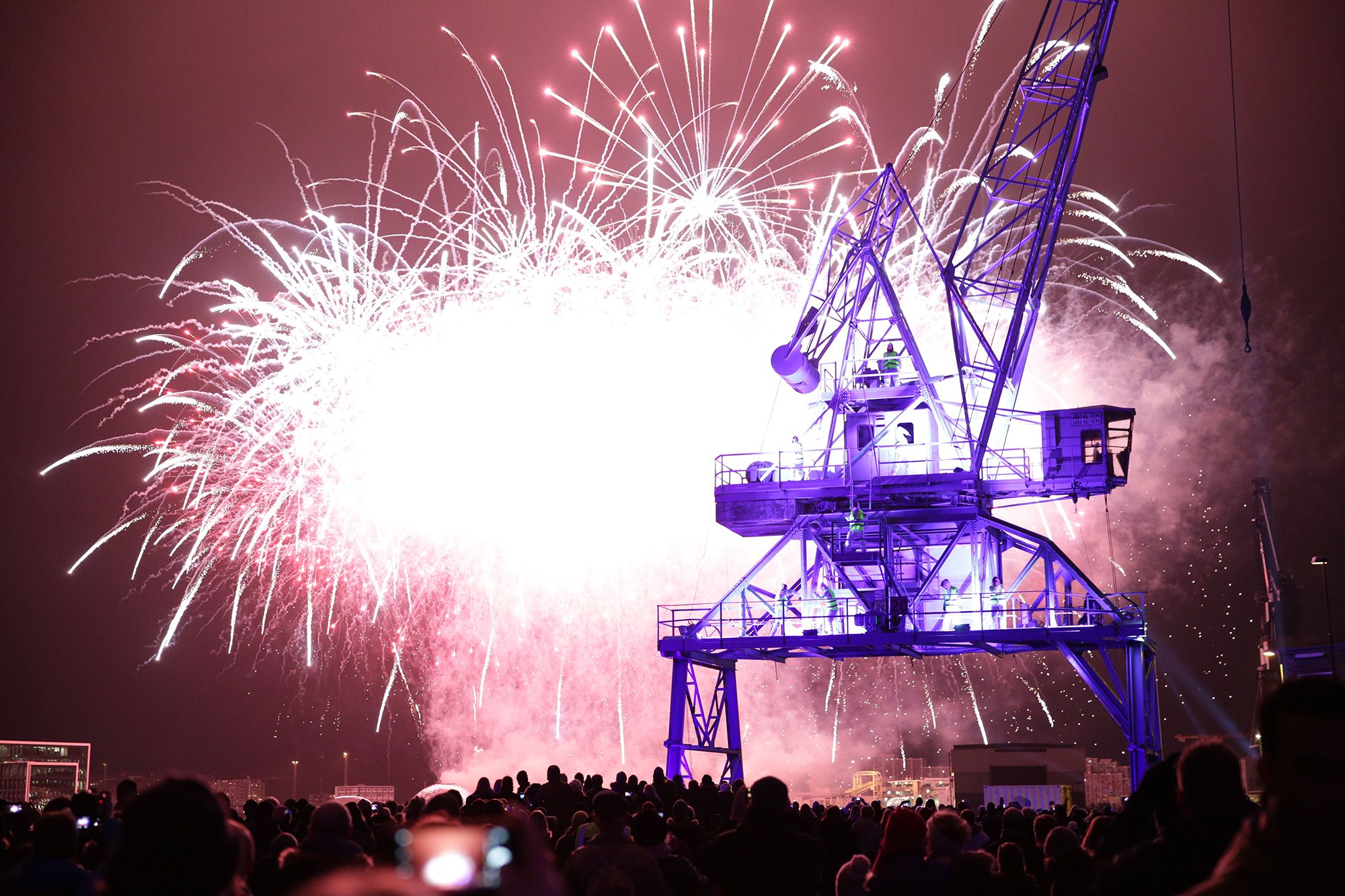 A burst of white fireworks behind the silhouette of a harbour crane, with a crowd watching in the foreground.